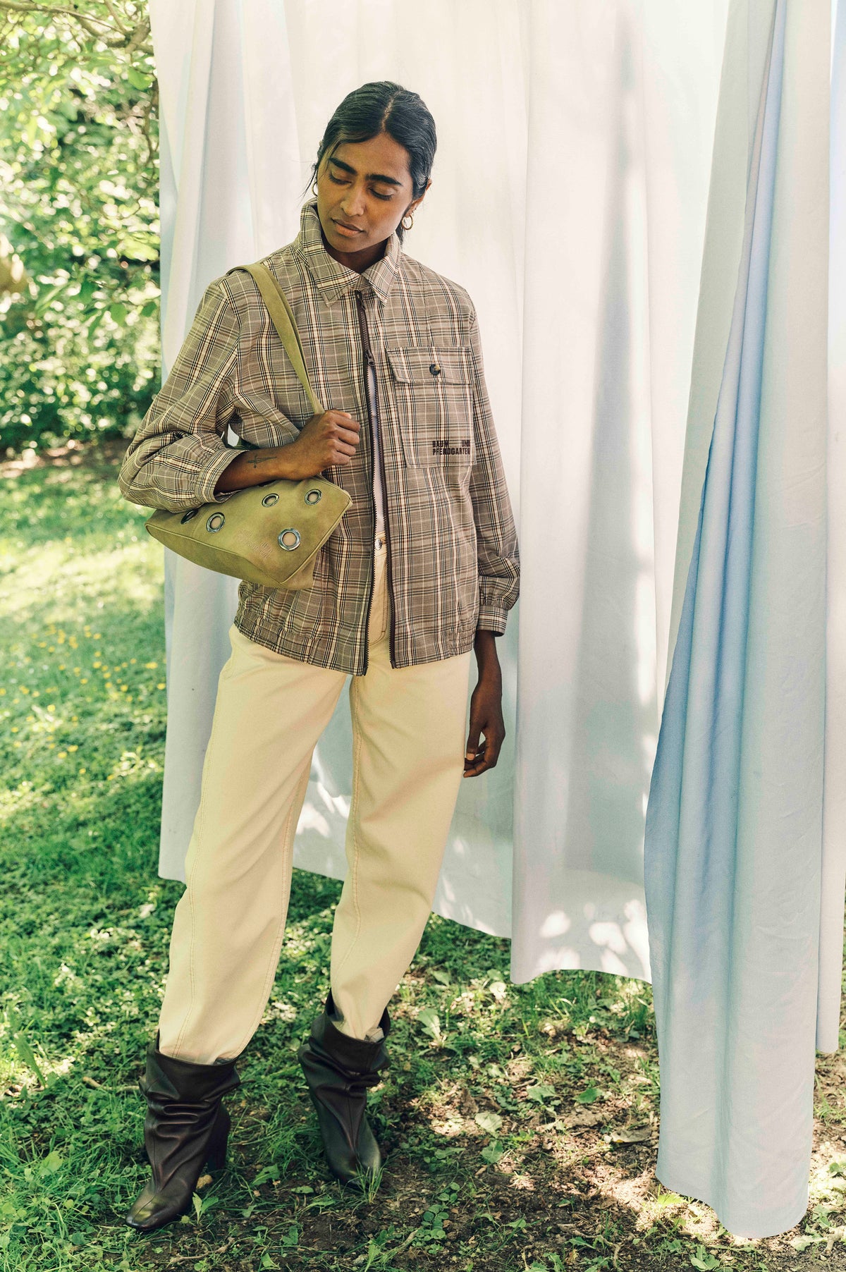 Campaign portrait of a model in a beige plaid zip-front jacket and cream trousers, carrying an olive bag beside sheer curtains outdoors.