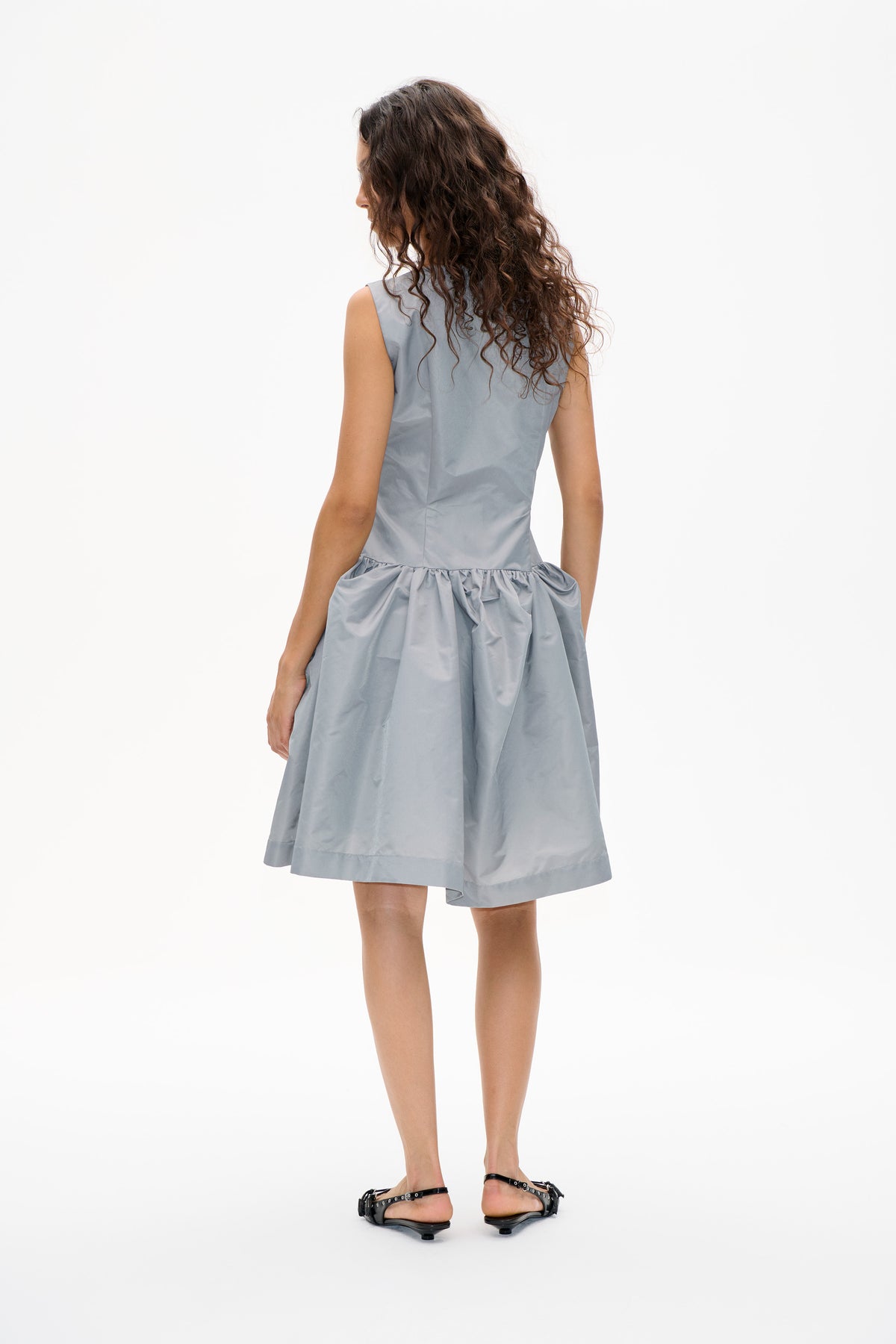 Back view of a model wearing a pale blue-gray sleeveless dress with a fitted bodice and gathered, voluminous skirt.