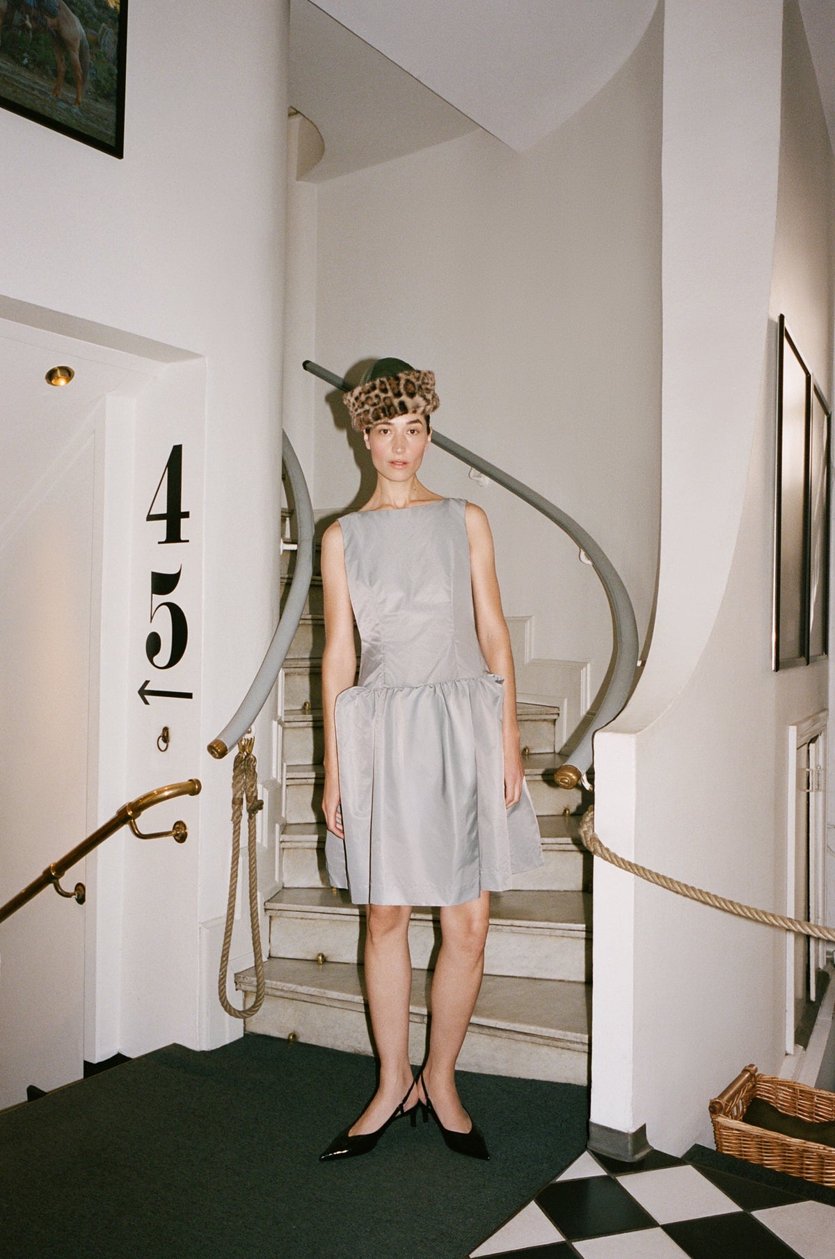 Model wearing a sleeveless pale grey dress with a gathered waist and full skirt, paired with a leopard-print fur hat and black pointed shoes on a staircase.