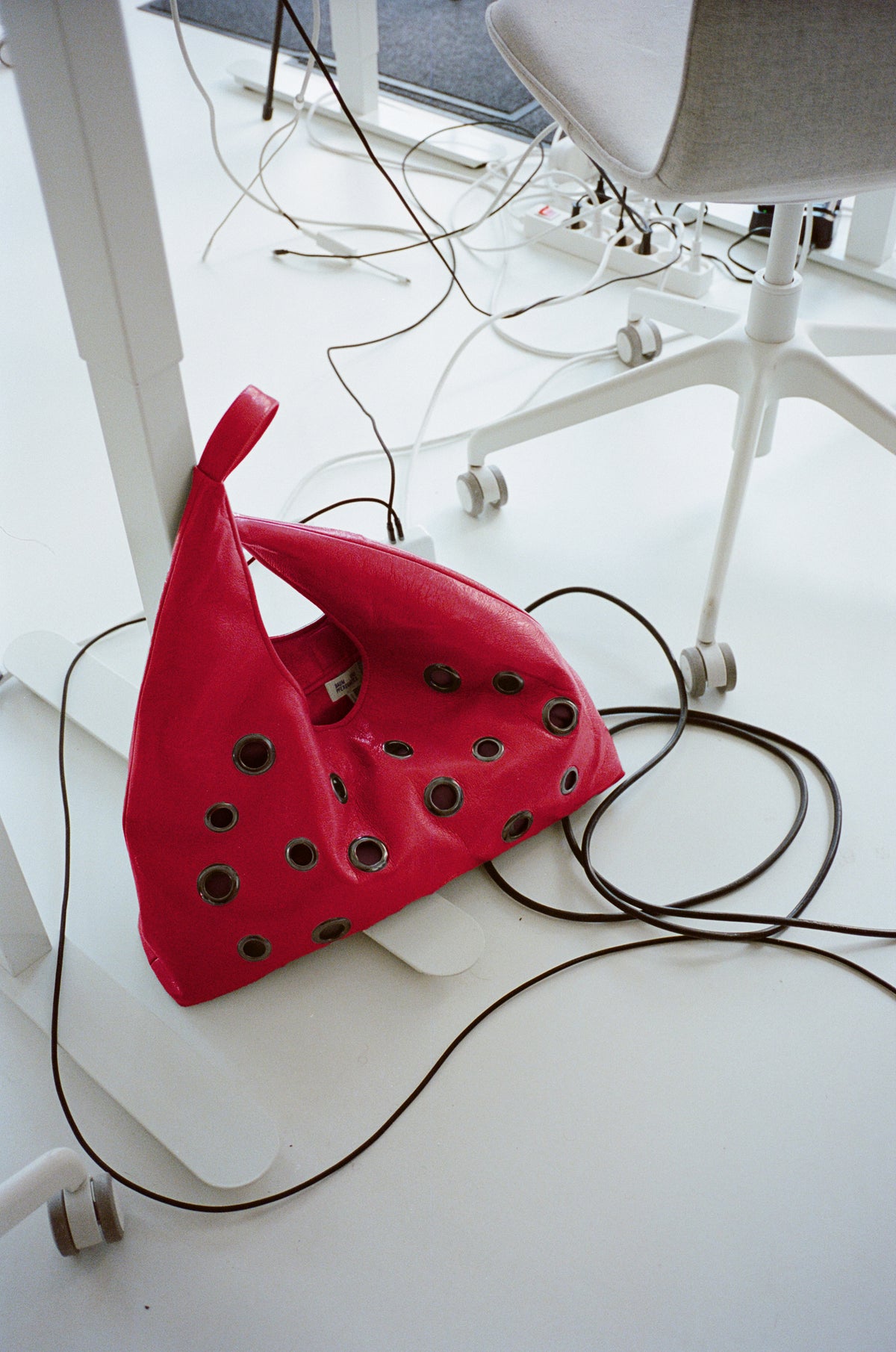 Bright red shoulder bag with oversized metal eyelets and long straps, styled on a white floor among chair legs and cables.