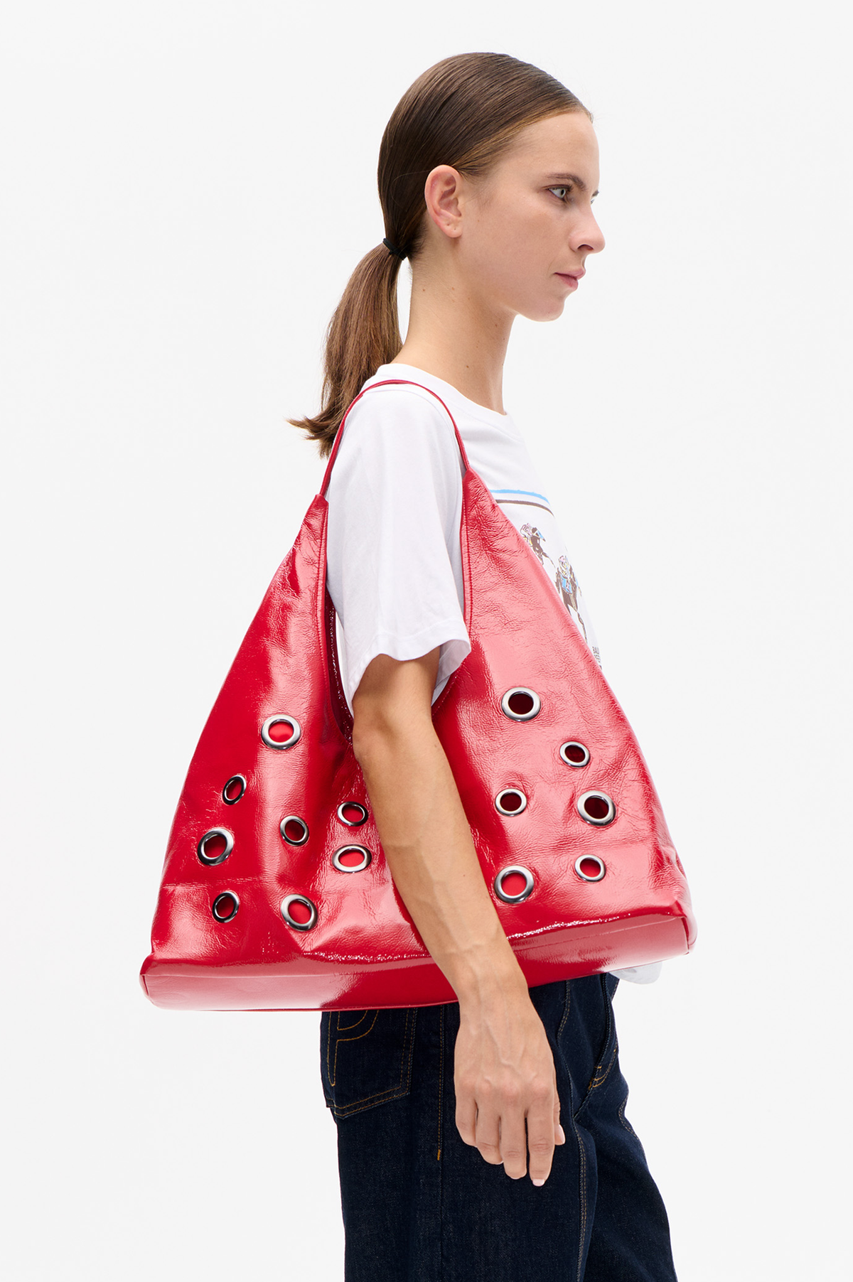 Side-profile campaign portrait of a model carrying a glossy red oversized tote with silver eyelets, wearing a white graphic T-shirt and dark jeans.