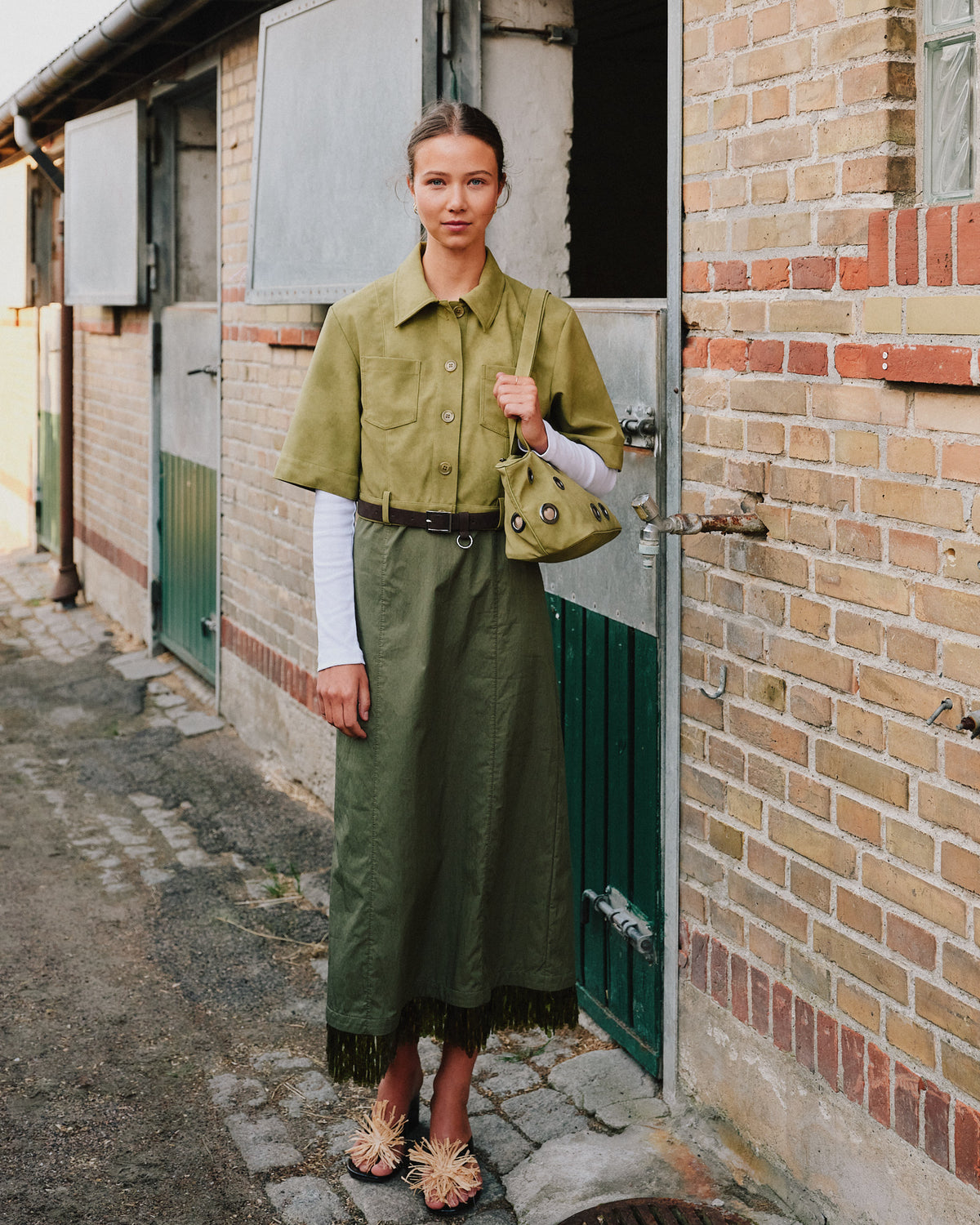 Model wearing an olive green button-front shirt dress with chest pockets and a belt, carrying a matching eyelet bag outside a brick building.