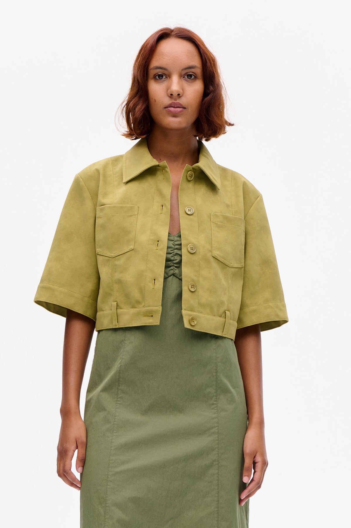 Front view of a model wearing a yellow-olive cropped jacket with short sleeves, a wide collar, button front and large patch pockets over a green dress.