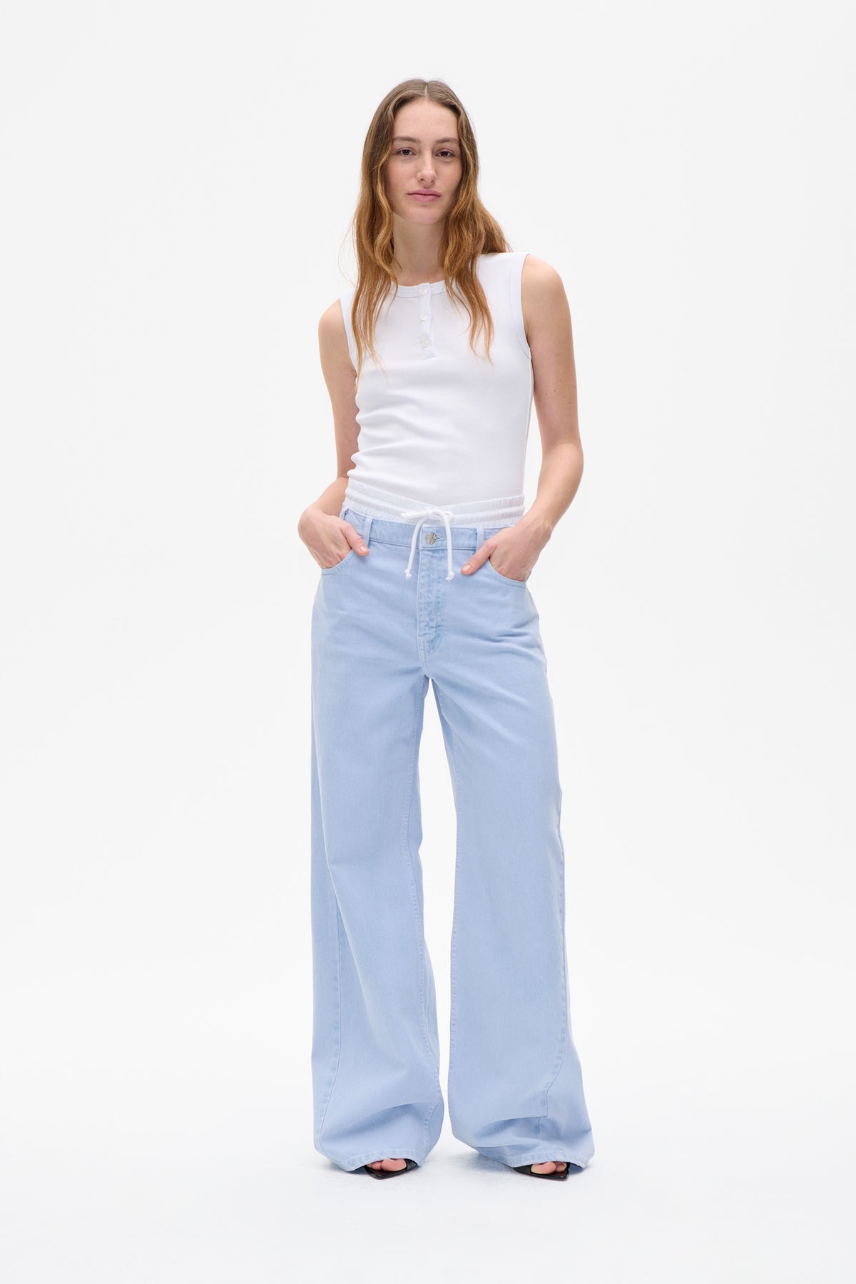 Model trägt ein weißes, ärmelloses Top mit Knopfleiste und hellblaue, weit geschnittene Jeans mit gerafftem Kordelzugbund, von vorne.