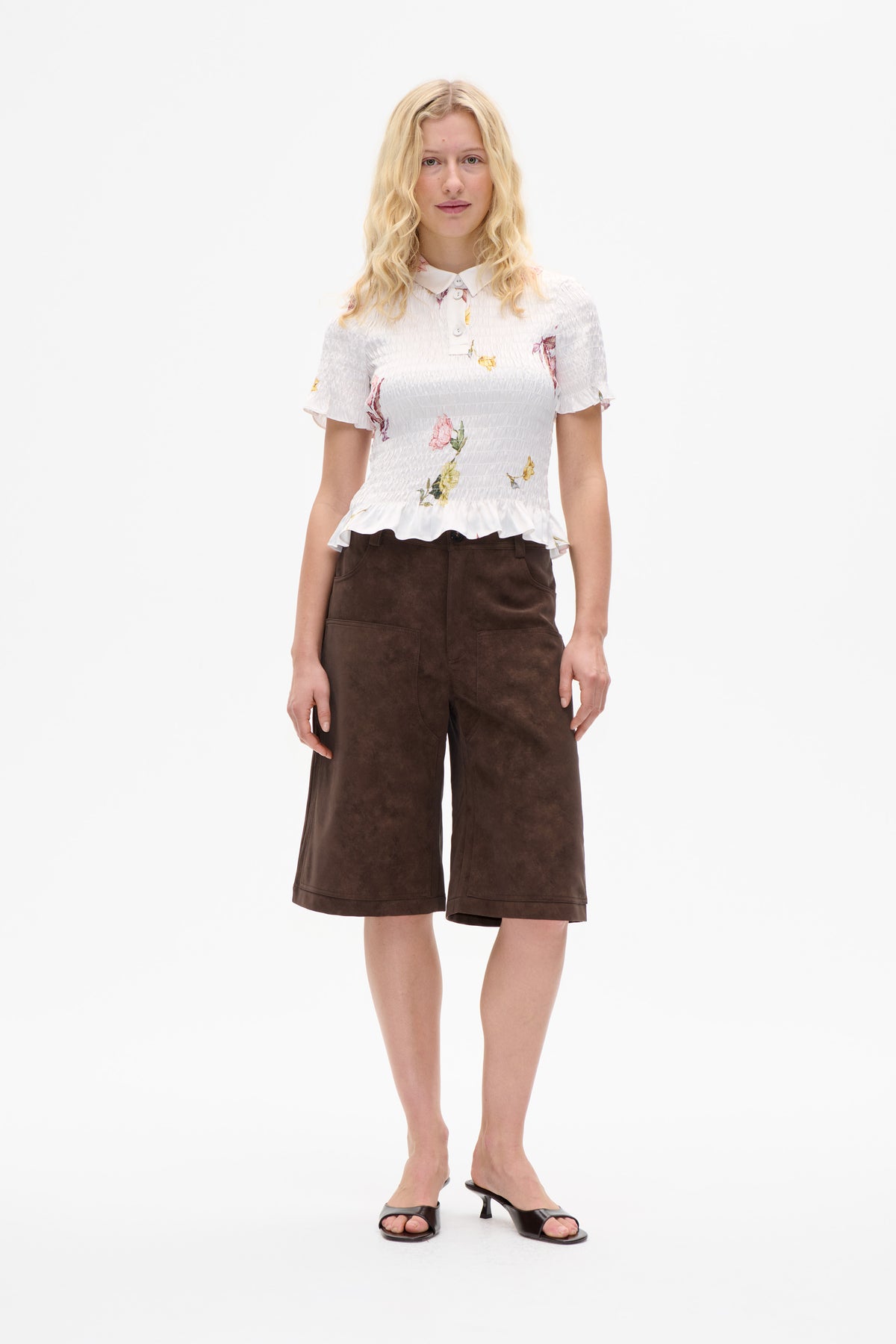 Model wearing a white floral-print smocked blouse with a pointed collar and ruffled hem, paired with brown wide-leg shorts.