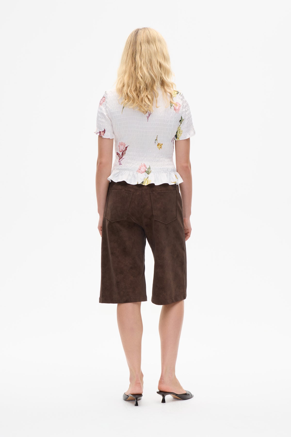 Back view of a model wearing a white smocked floral blouse with ruffled sleeves and hem, brown wide-leg cropped trousers, and black heeled mules.