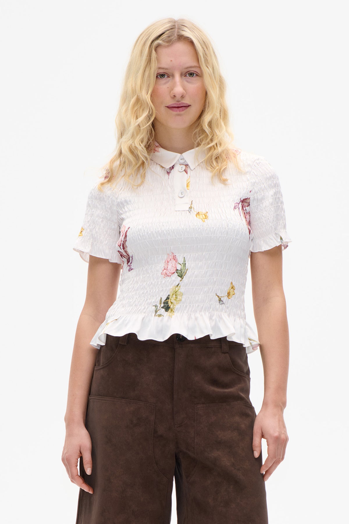 Front view of a model wearing a white floral-print blouse with a pointed collar, smocked bodice and flounced sleeves, paired with brown trousers.