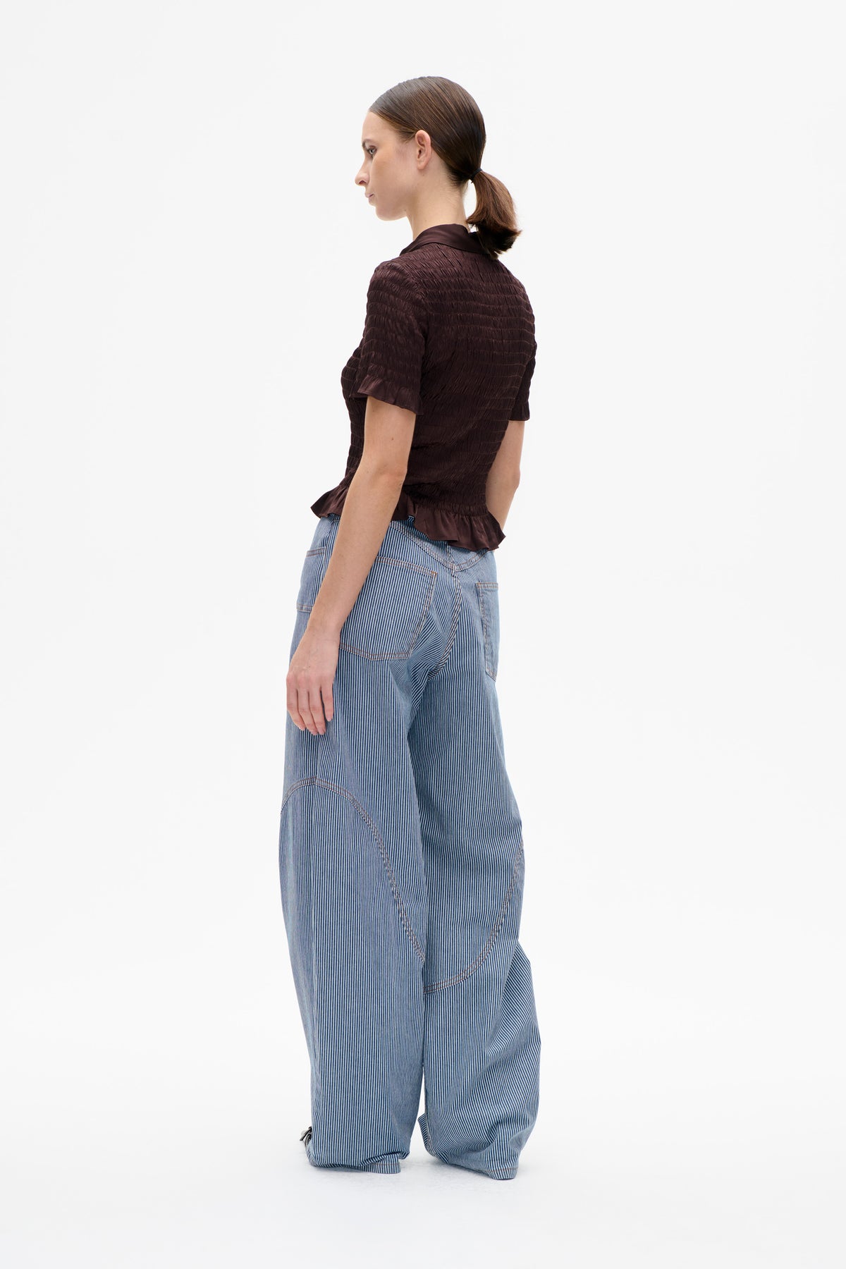 Model shown from the side and back wearing a dark plum ruched short-sleeve collared blouse with a flared hem and blue pinstriped trousers.