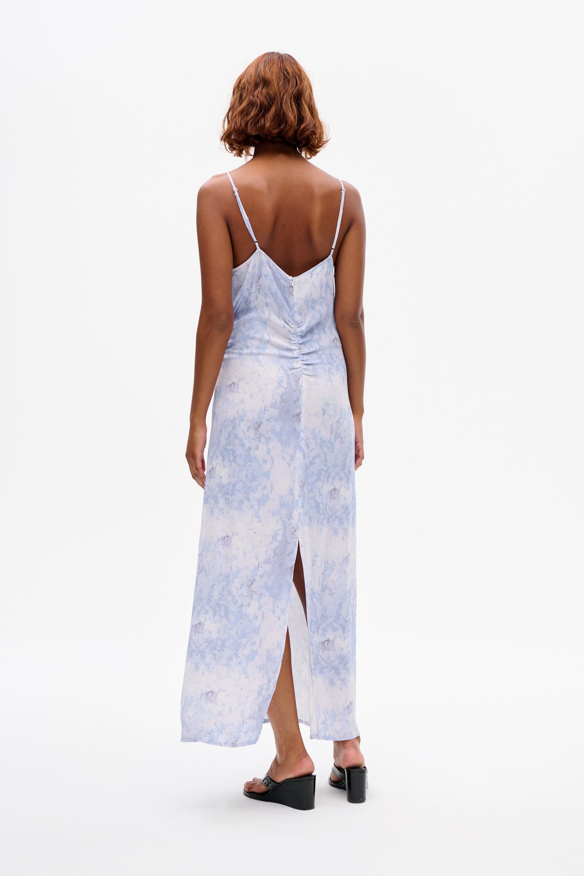 Back view of a model wearing a pale blue and white floral-print spaghetti-strap maxi dress with a low back and side slit.