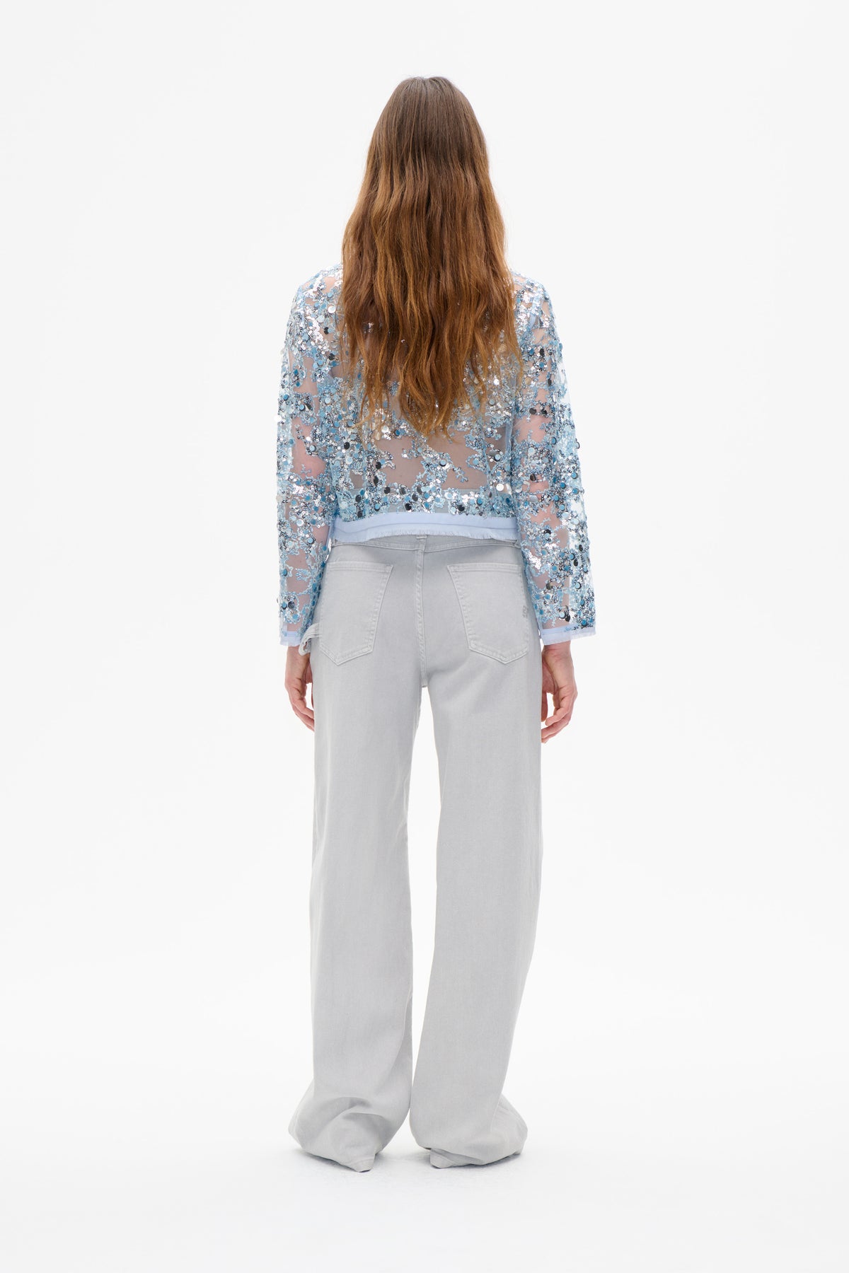 Achteraanzicht van een doorschijnende lichtblauwe top met lange mouwen, geheel bedekt met zilveren en blauwe pailletten, gecombineerd met lichtgrijze jeans met wijde pijpen.