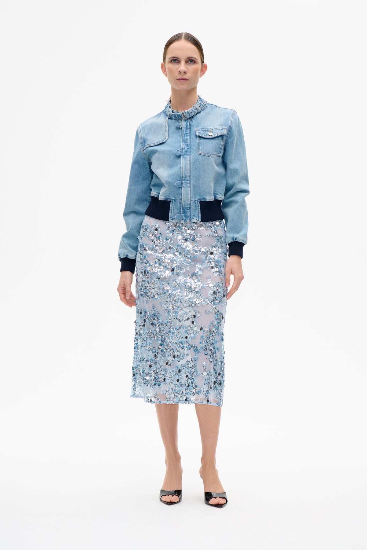 Front view of a model wearing a light blue denim jacket with a buckle collar and navy ribbed trim, paired with a silver-blue sequin skirt.