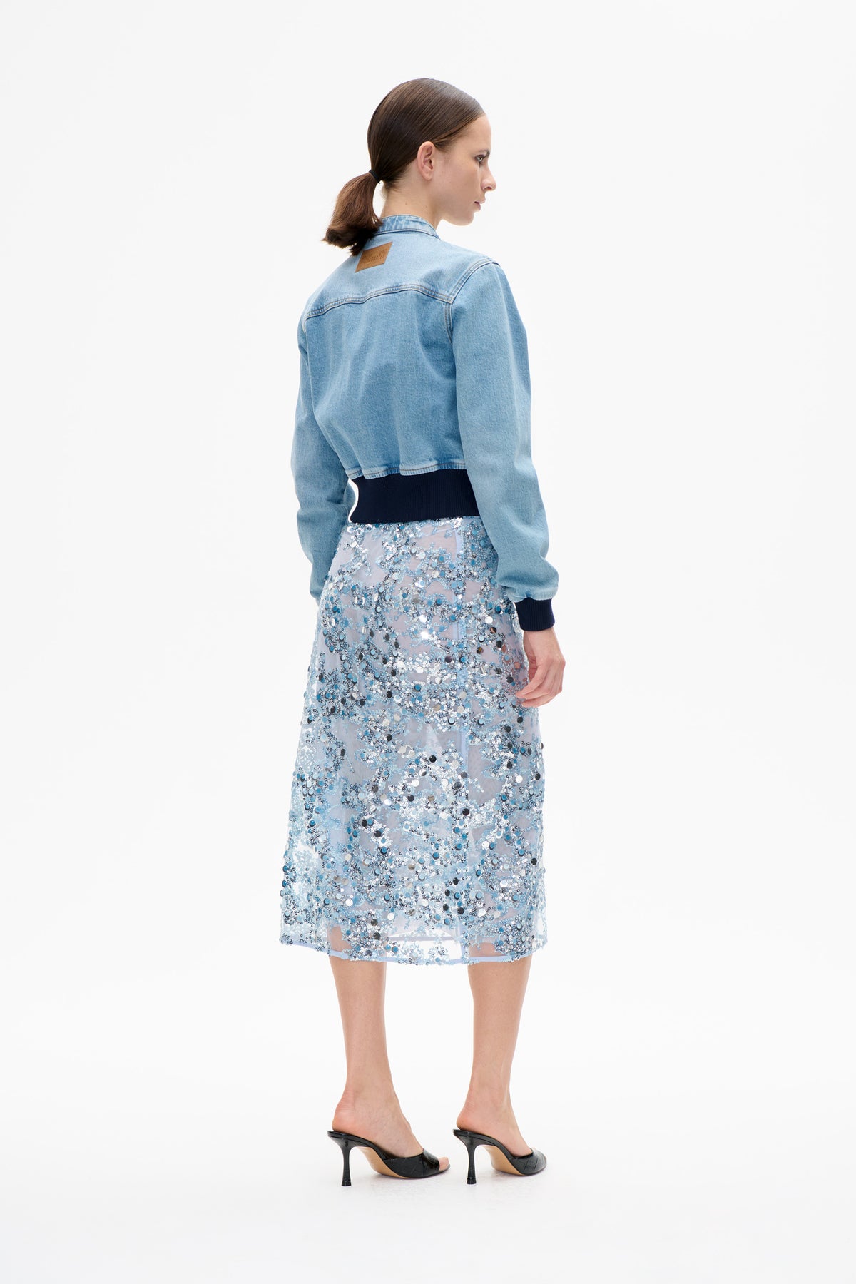 Model wearing a light blue denim jacket with dark ribbed cuffs, shown from the back with a pale blue and silver sequin midi skirt.