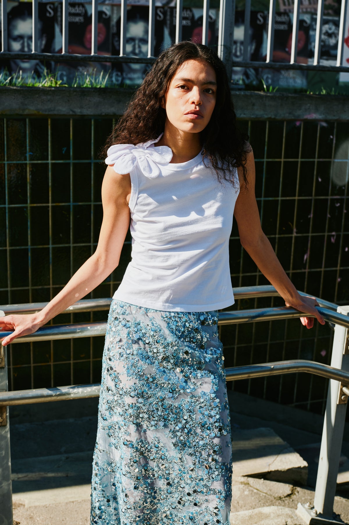 Campagneportret van een model dat een witte mouwloze top met een stoffen bloem op de schouder en een blauwe paillettenrok draagt.