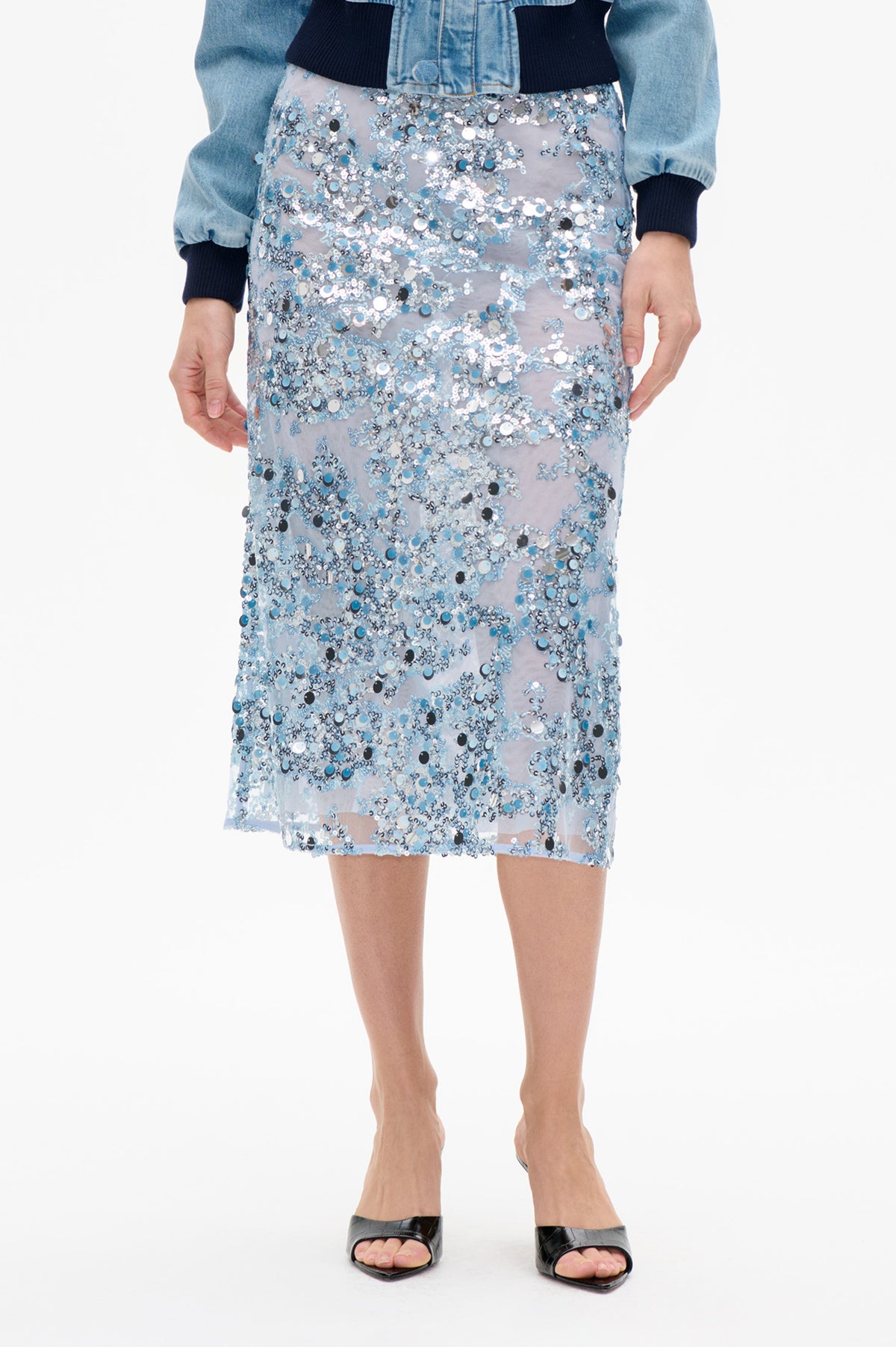 Detail van de voorkant van een doorschijnende, lichtblauwe knielange rok, dicht bezet met blauwe, zilveren en zwarte pailletten, gestyled met een licht denimjack.