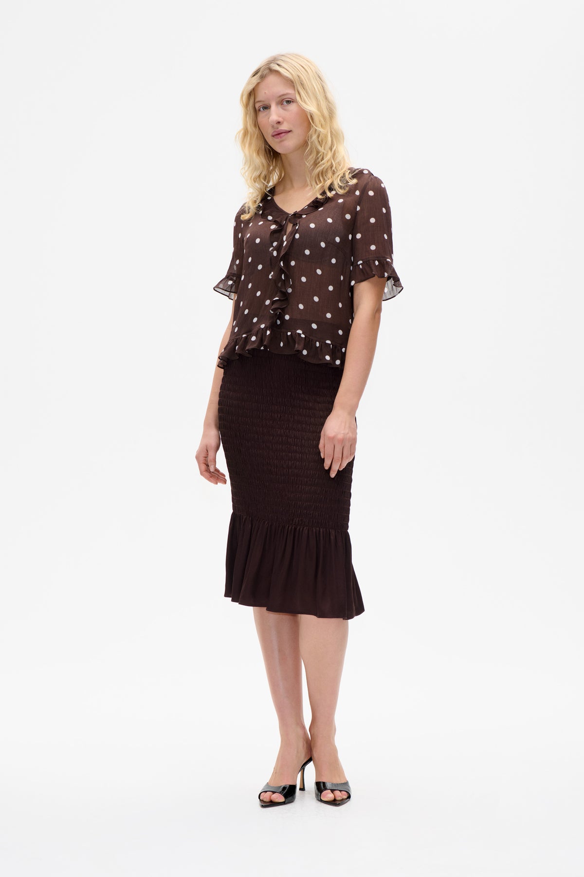 Front view of a model wearing a brown blouse with white polka dots, ruffled neckline and sleeves, paired with a dark smocked skirt.