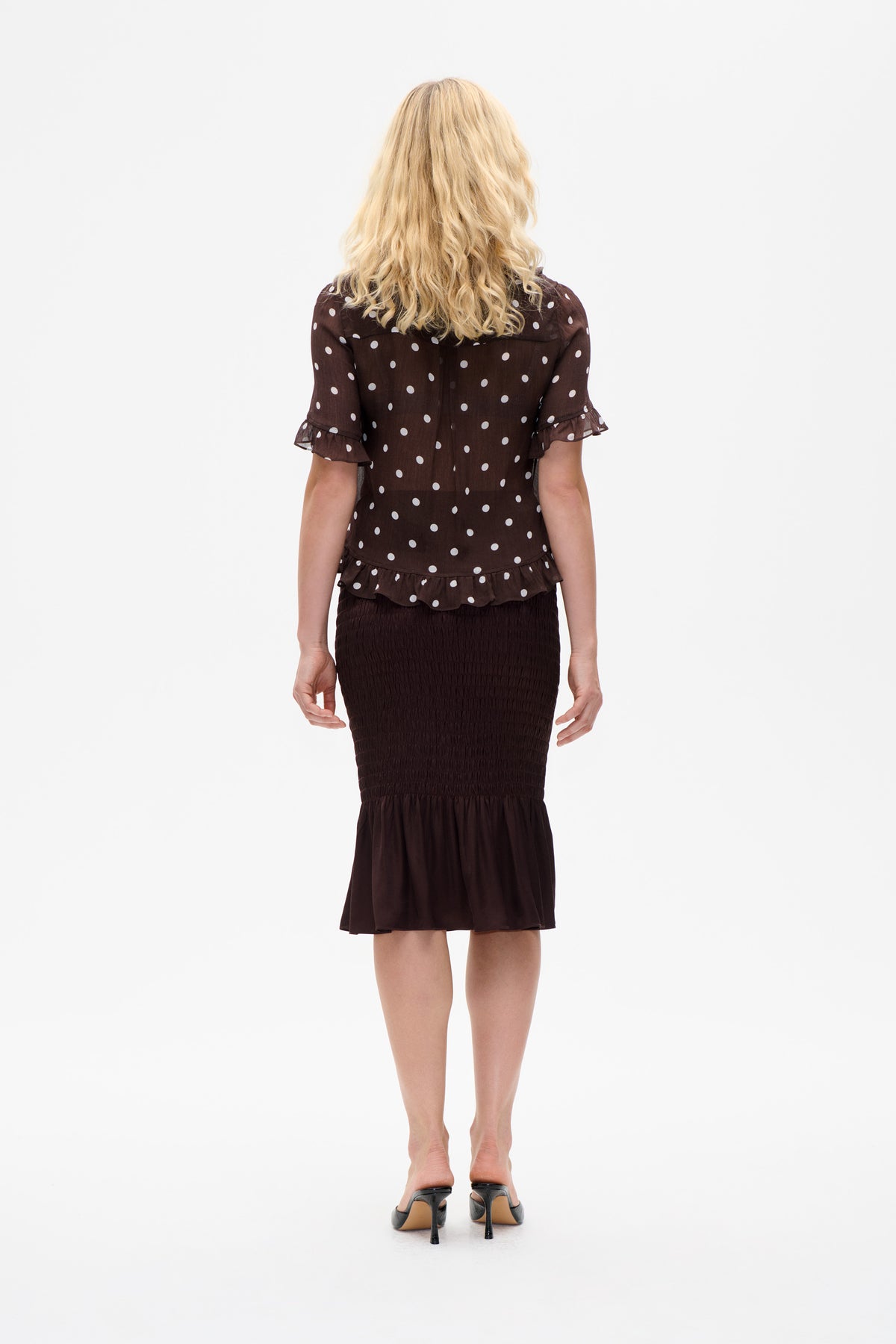 Back view of a dark brown blouse with white polka dots, flutter sleeves and a ruffled hem, paired with a dark ruched skirt.