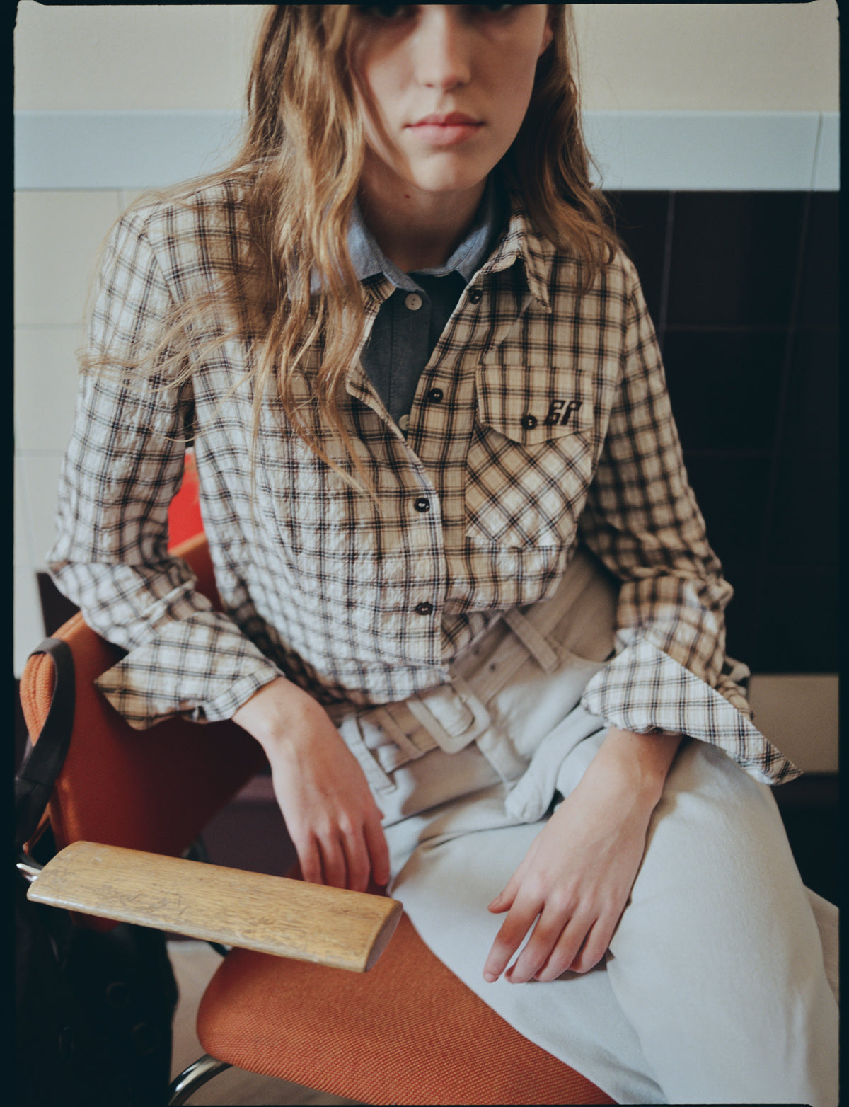 Zittend model draagt een beige-bruine geruite blouse met knopen over een donkere top met kraag en een lichte broek.