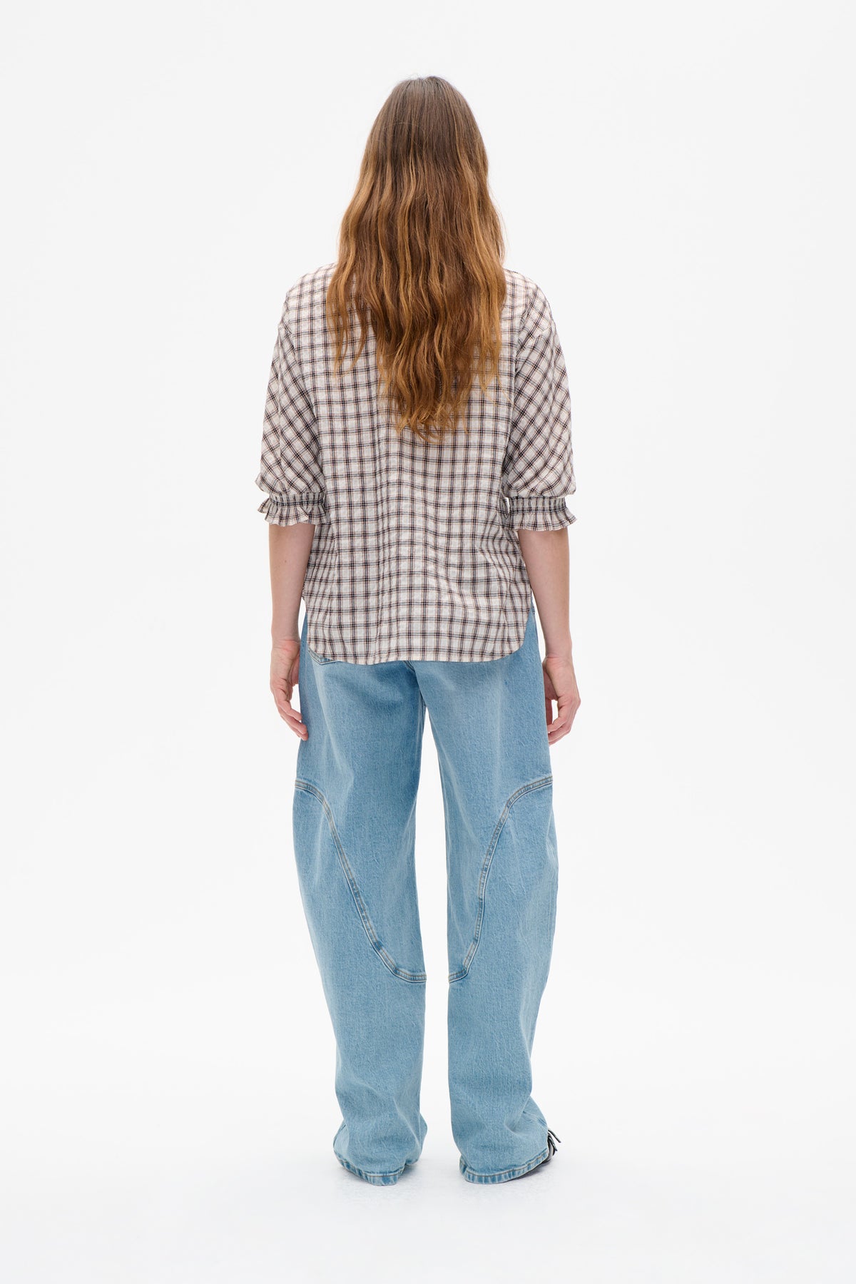 Achteraanzicht van een model met een crèmekleurige geruite blouse met geplooide ruches aan de manchetten en lichtblauwe jeans met wijde pijpen.