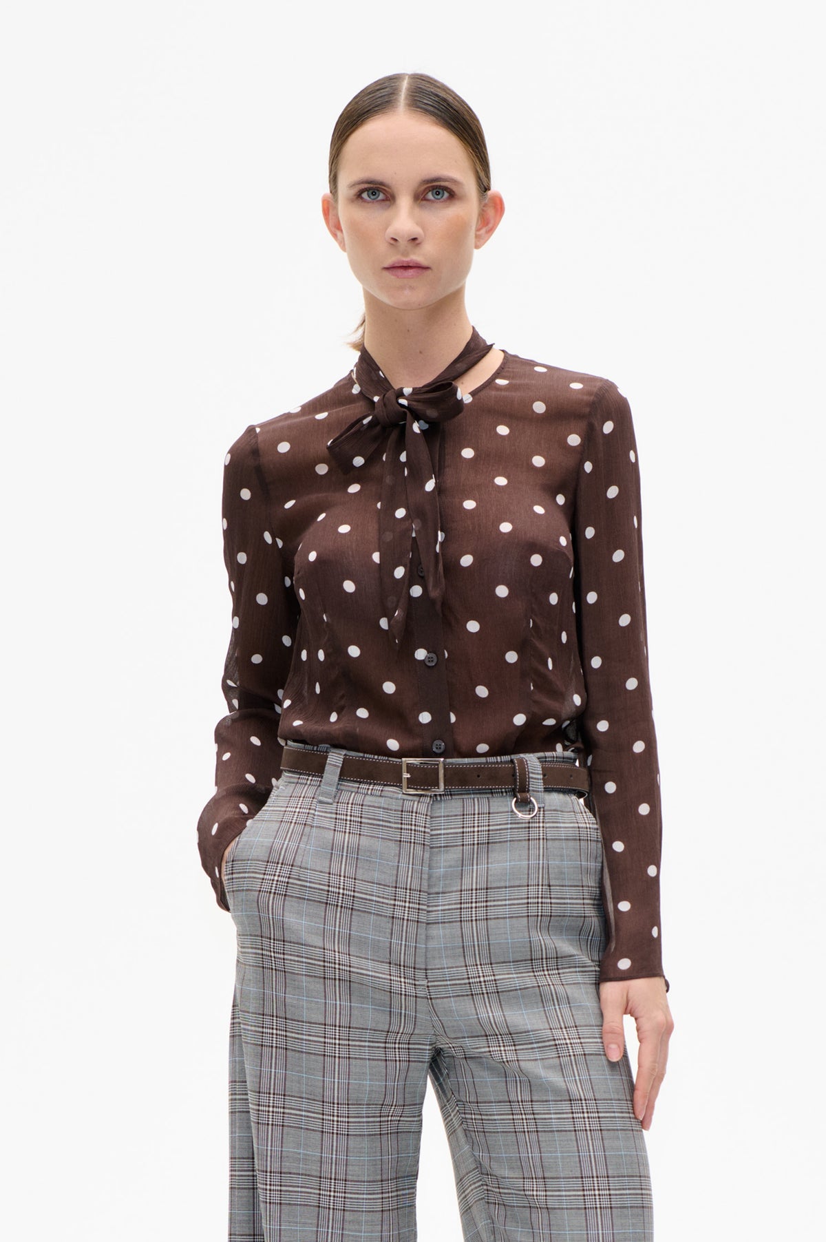 Front view of a brown blouse with white polka dots and a tied neck scarf, paired with grey plaid trousers and a brown belt.