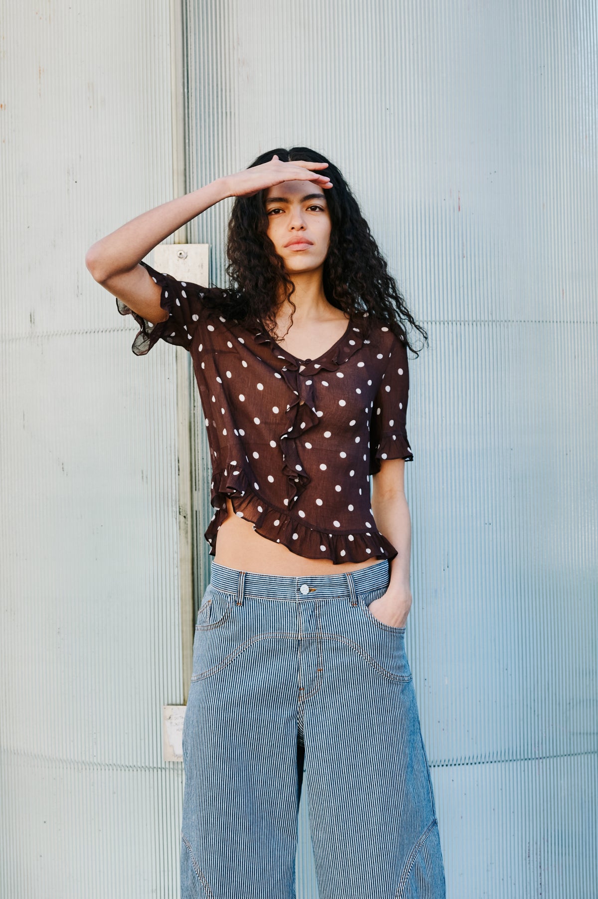 Model poses with one hand shading her eyes, wearing a dark brown blouse with white polka dots and ruffles, and blue-and-white striped wide-leg jeans.