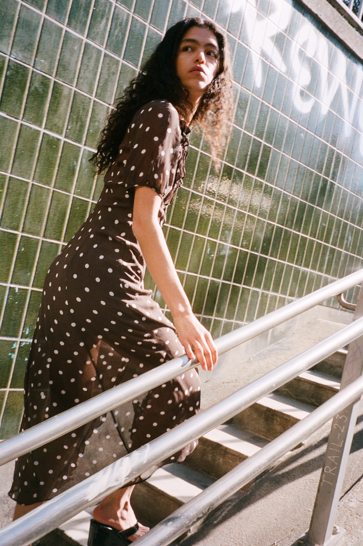 Model in a black short-sleeved dress with white polka dots, climbing stairs beside a tiled wall