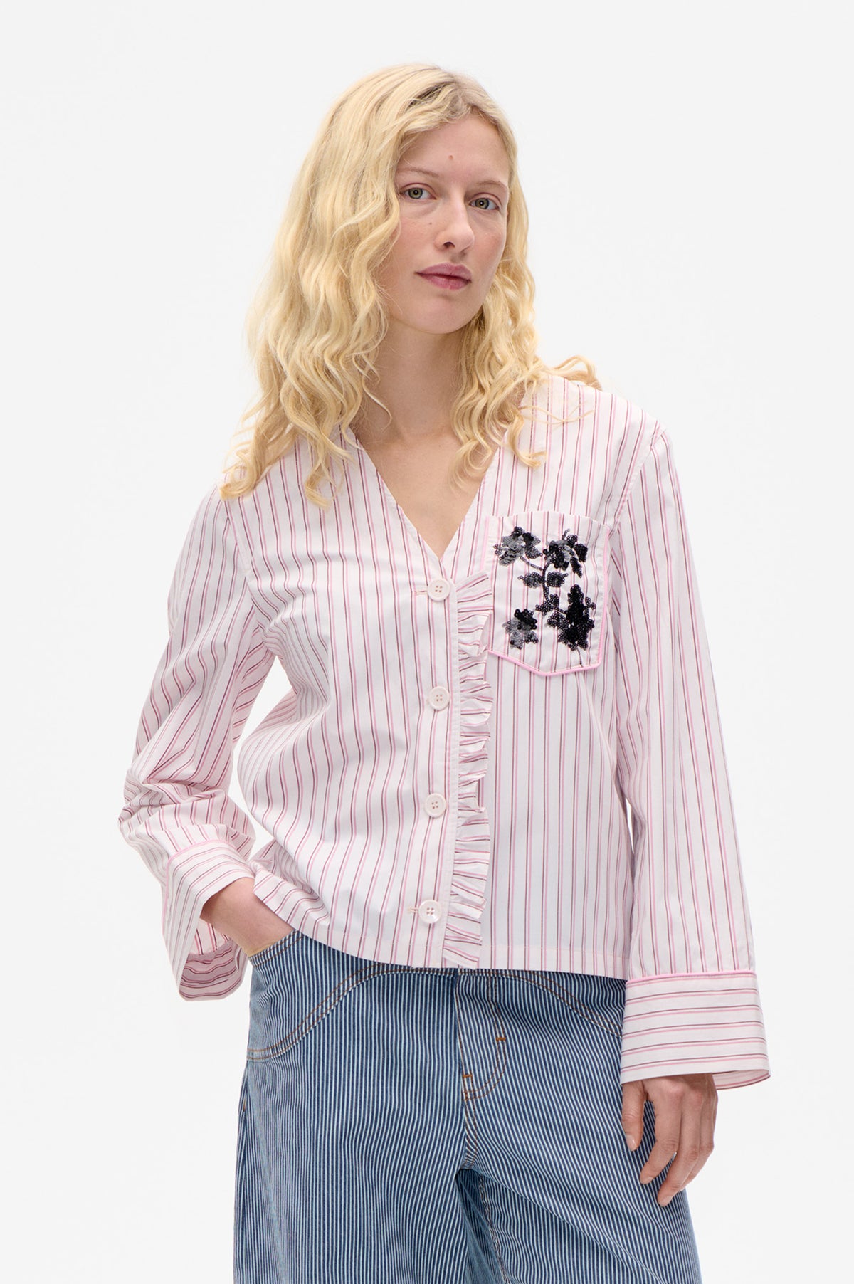 Model wearing a white shirt with pink and grey stripes, a ruffled button placket and black floral embellishment, paired with blue pinstripe trousers.