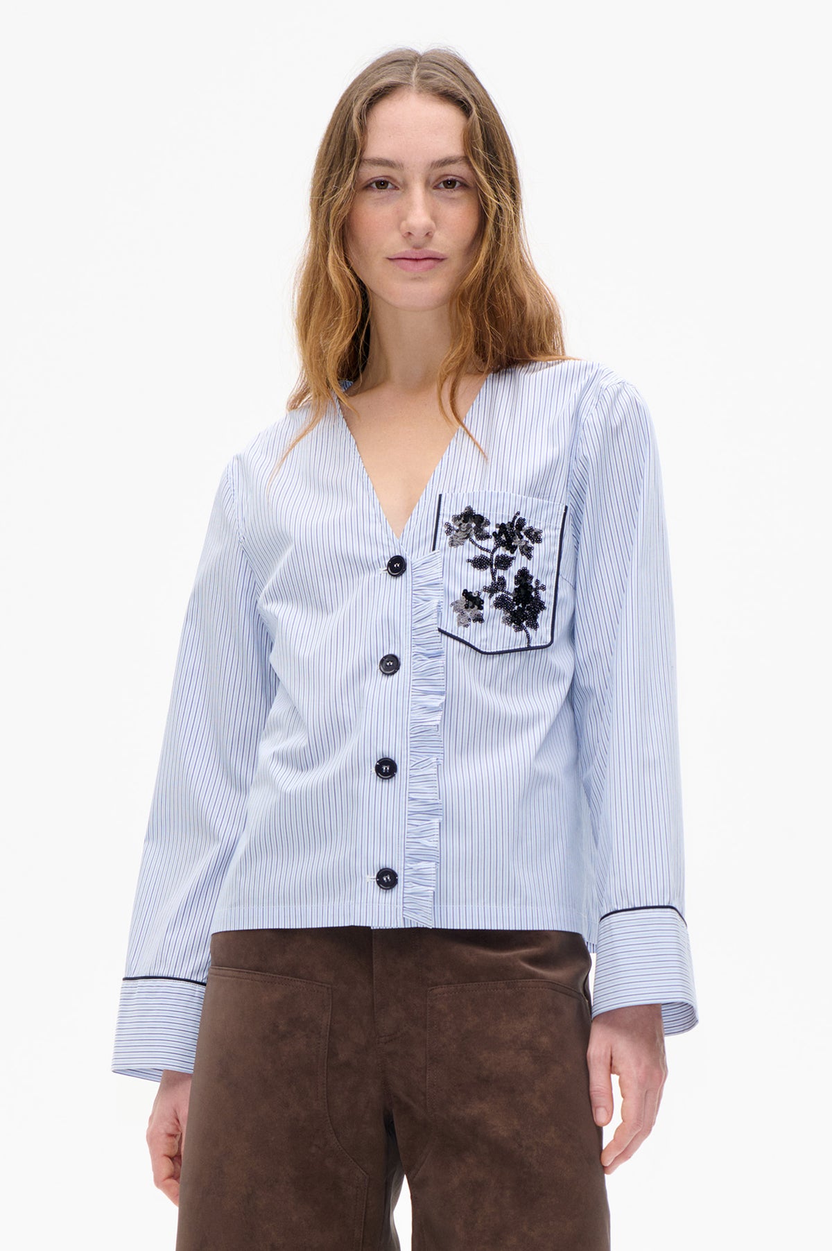 Model wearing a white blouse with fine blue stripes, black sequin floral pocket, contrast piping and dark buttons, paired with brown trousers.