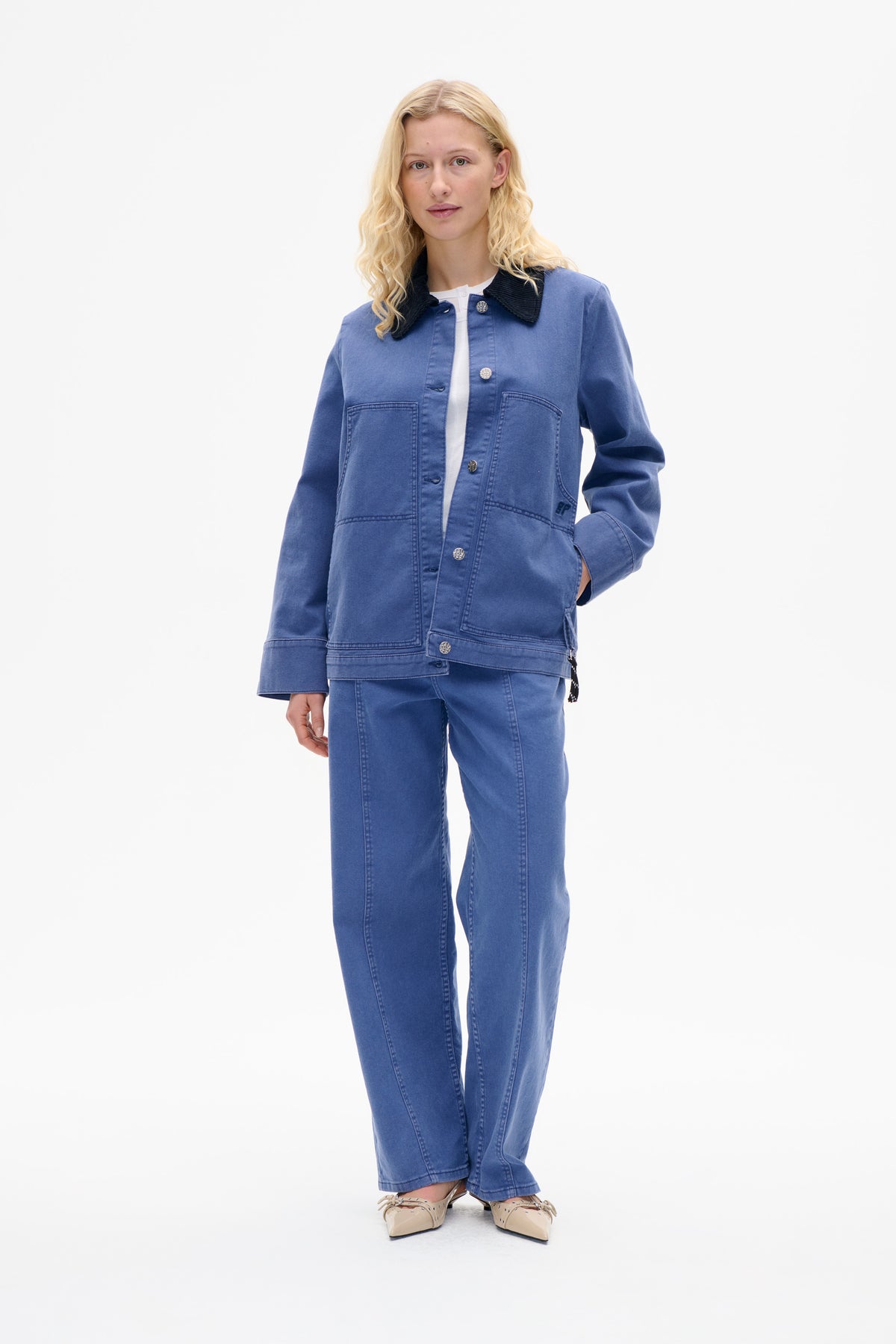 Model wearing a blue button-front jacket with a black collar and large patch pockets, paired with matching blue trousers; front view.
