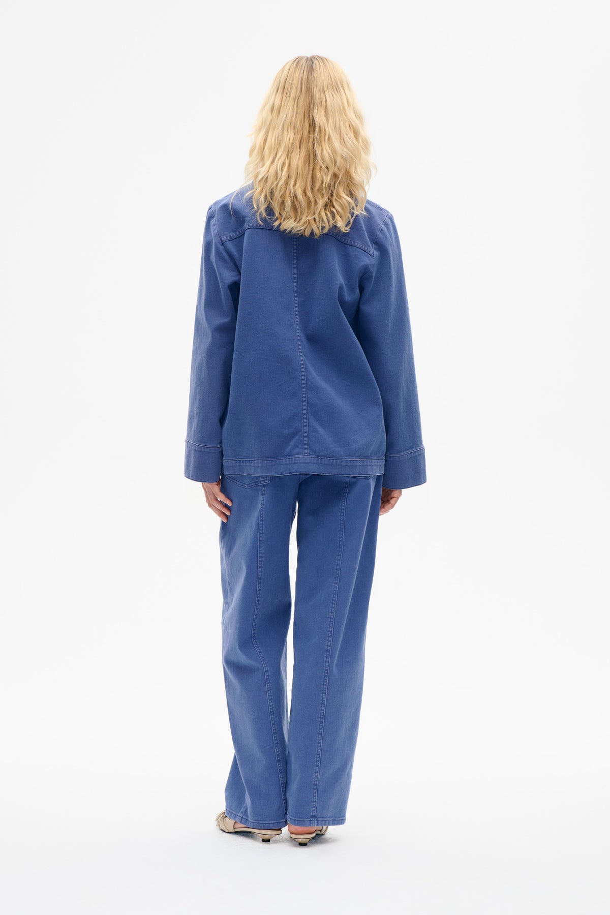 Back view of a model wearing a blue denim jacket with panel seams and matching straight-leg jeans.
