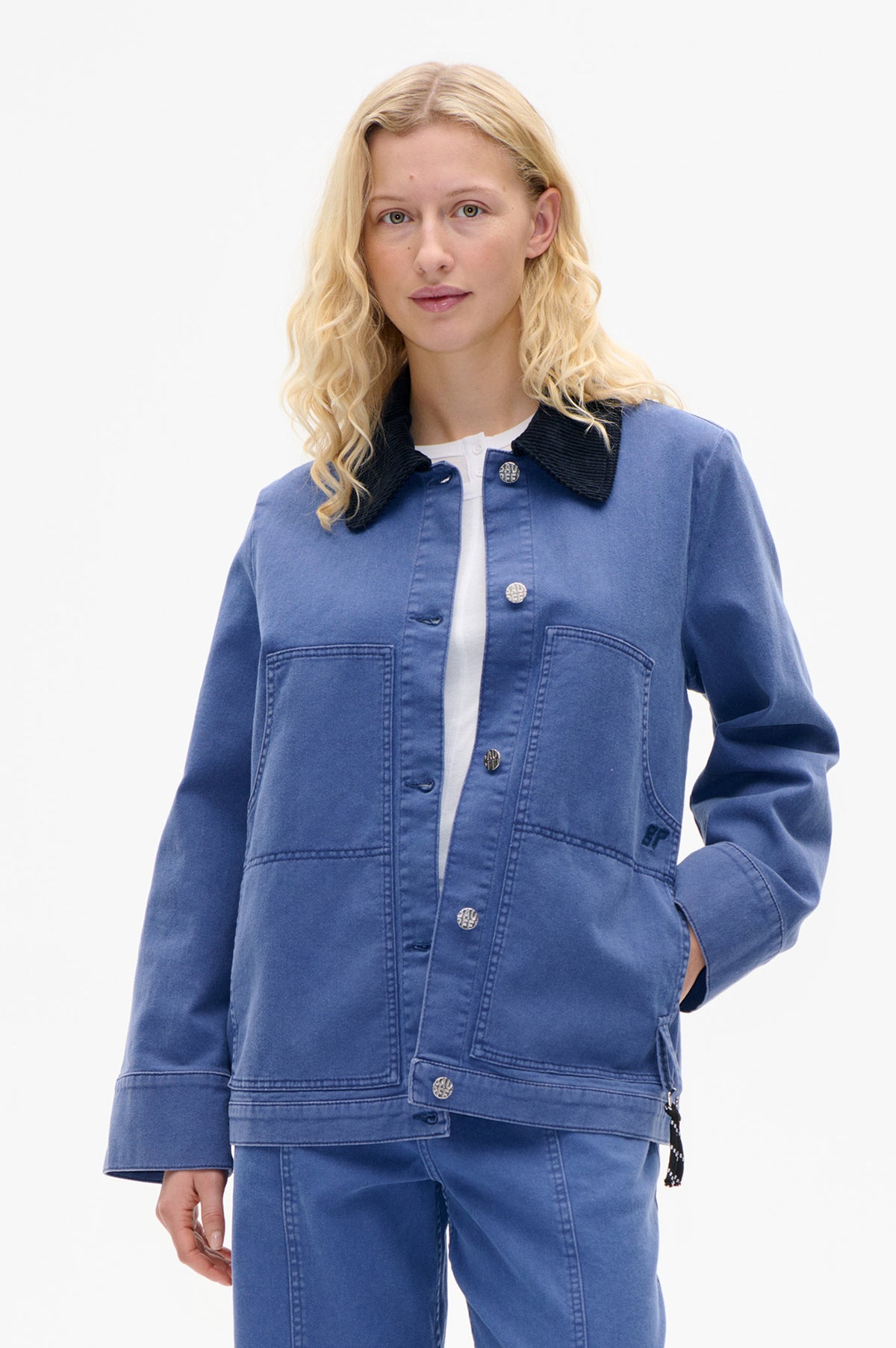 Model wearing a blue utility jacket with a black ribbed collar, silver buttons and large front pockets, styled with blue trousers.