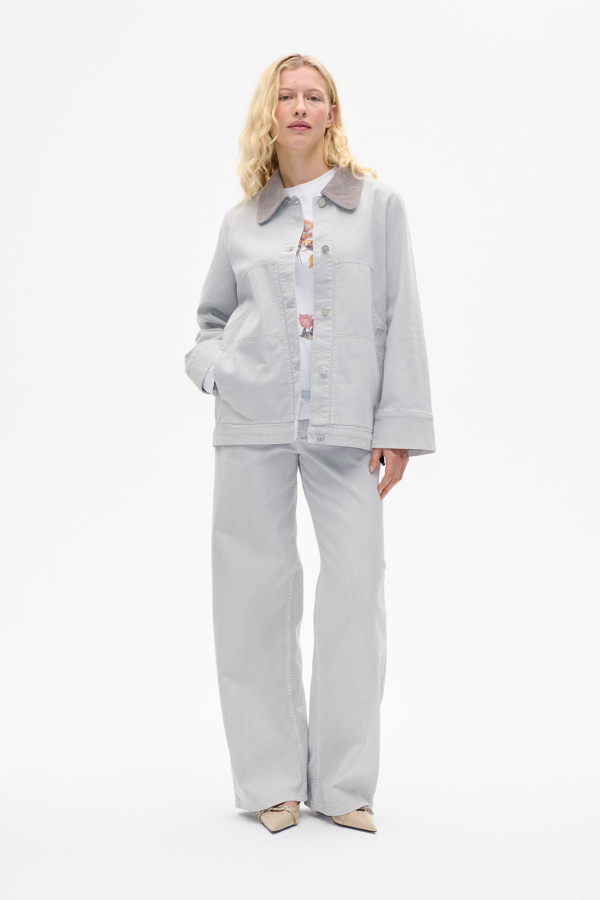 Model wearing a pale grey button-front jacket with a ribbed contrasting collar and large patch pockets, paired with wide-leg trousers.