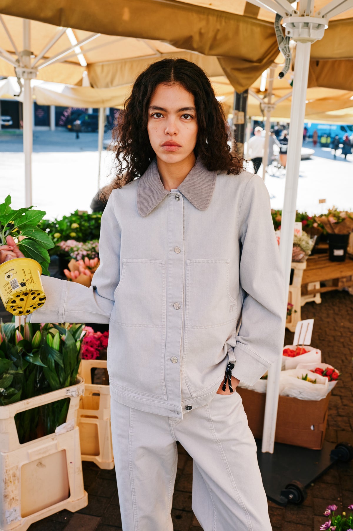Model wearing a pale blue-grey button-front jacket with a wide ribbed collar and patch pockets, paired with matching trousers at a flower stall.