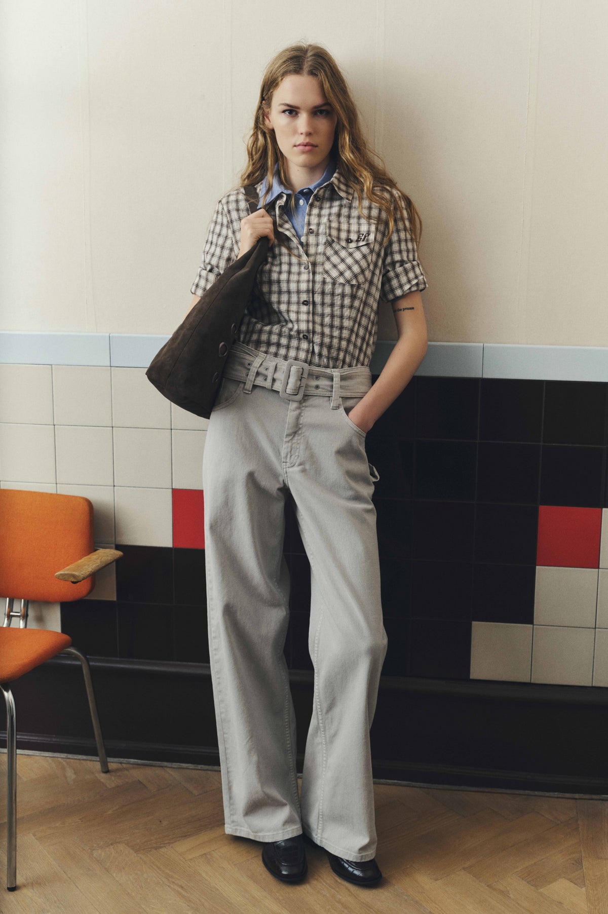 Model wearing a beige and black plaid short-sleeve shirt over a blue collared top with belted light-grey wide-leg jeans and a brown tote.
