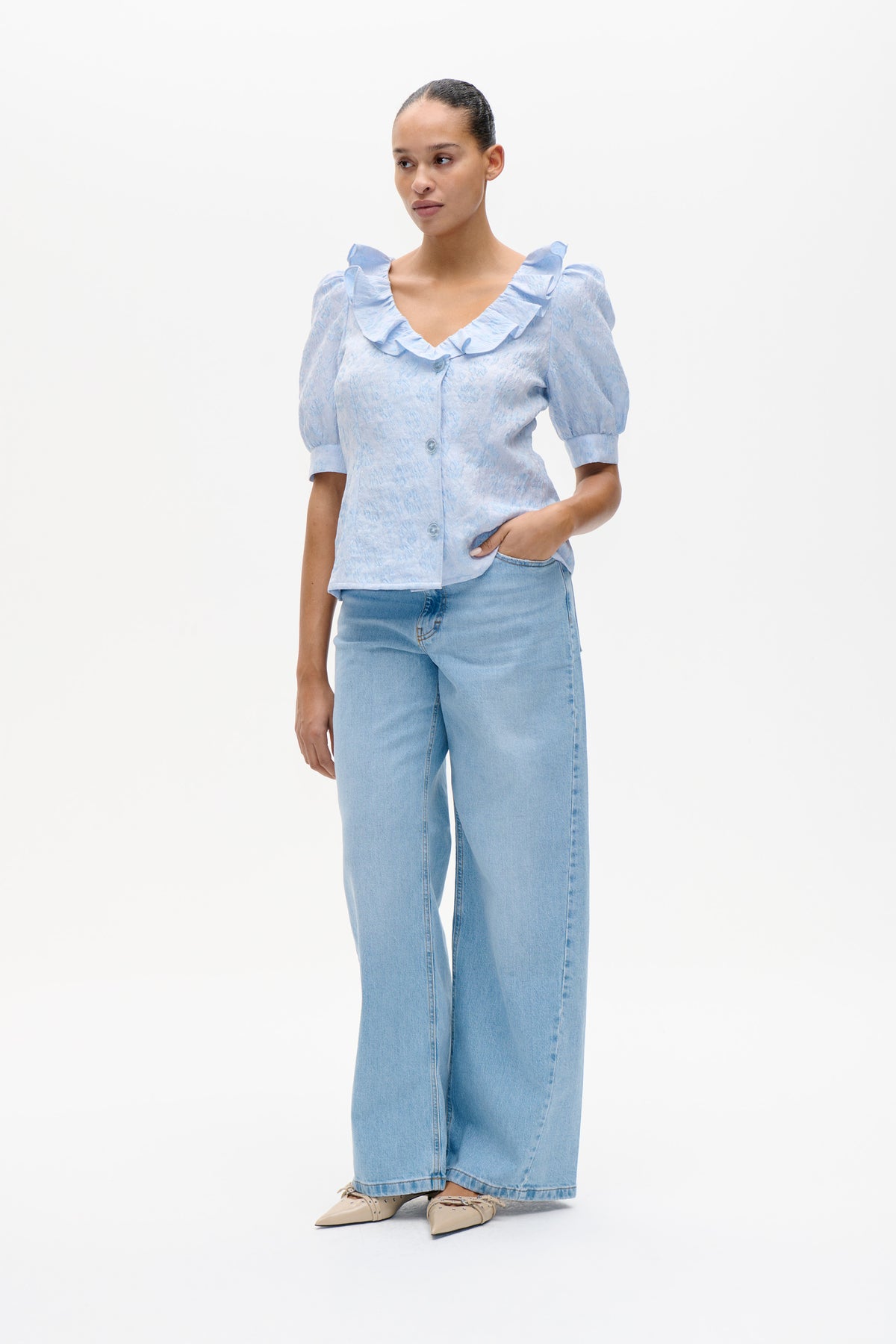 Model wearing a pale blue patterned blouse with a ruffled V-neck, button front and puff sleeves, paired with wide-leg light-wash jeans.
