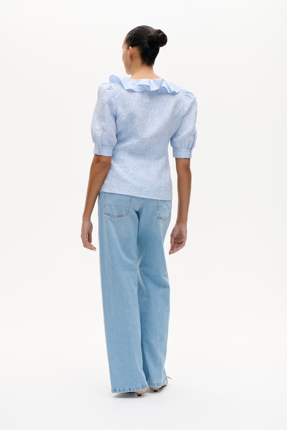 Back view of a pale blue blouse with a tonal textured pattern, ruffled collar and puff sleeves, paired with light-wash jeans.
