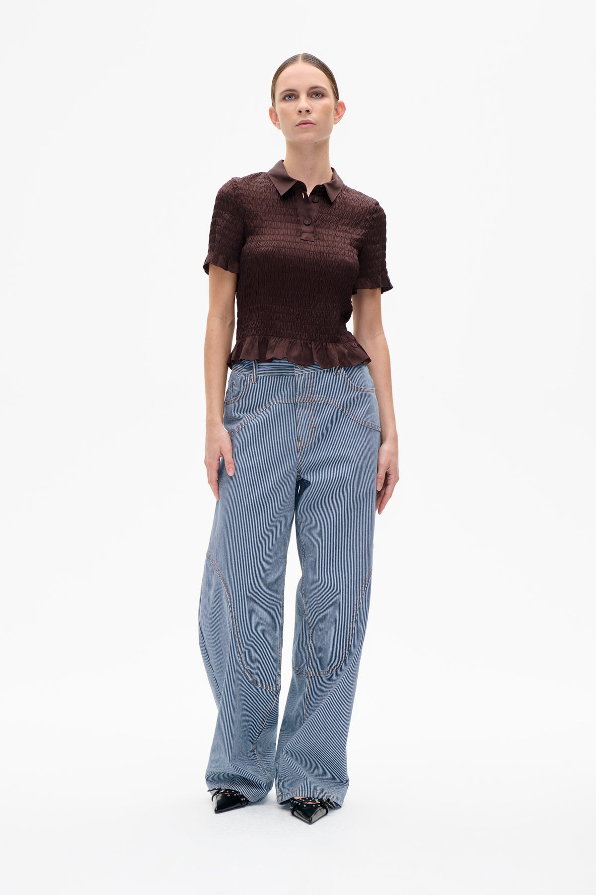 Front view of a model wearing a dark plum smocked short-sleeve collared top with a ruffled hem and blue pinstriped wide-leg trousers.