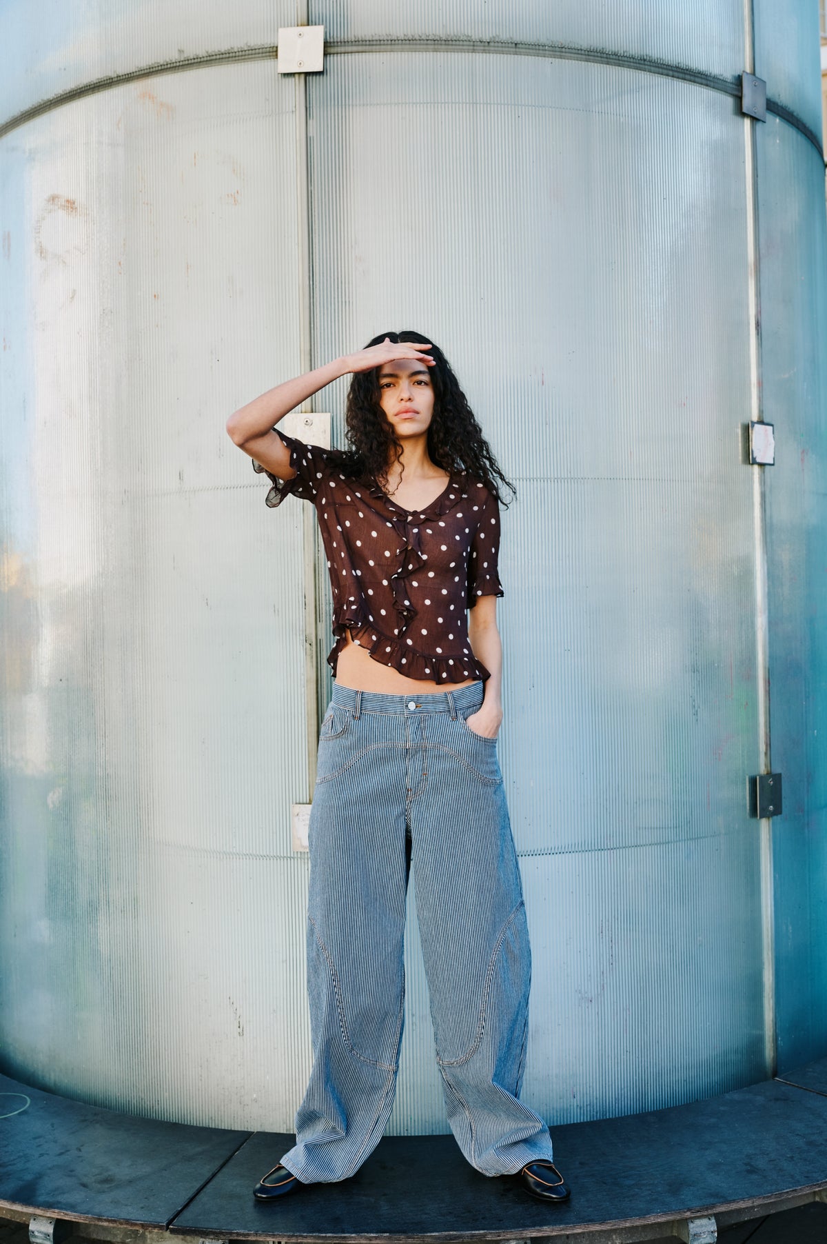 Campaign portrait of a model in a dark brown polka-dot ruffle blouse and wide-leg blue-and-white pinstriped trousers