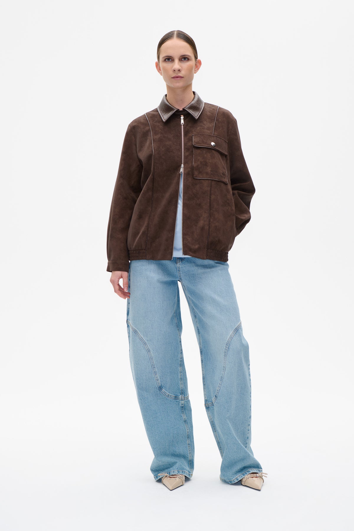 Front view of a model wearing a dark brown suede-look zip jacket with a contrasting pointed collar and flap chest pocket, paired with light blue jeans.