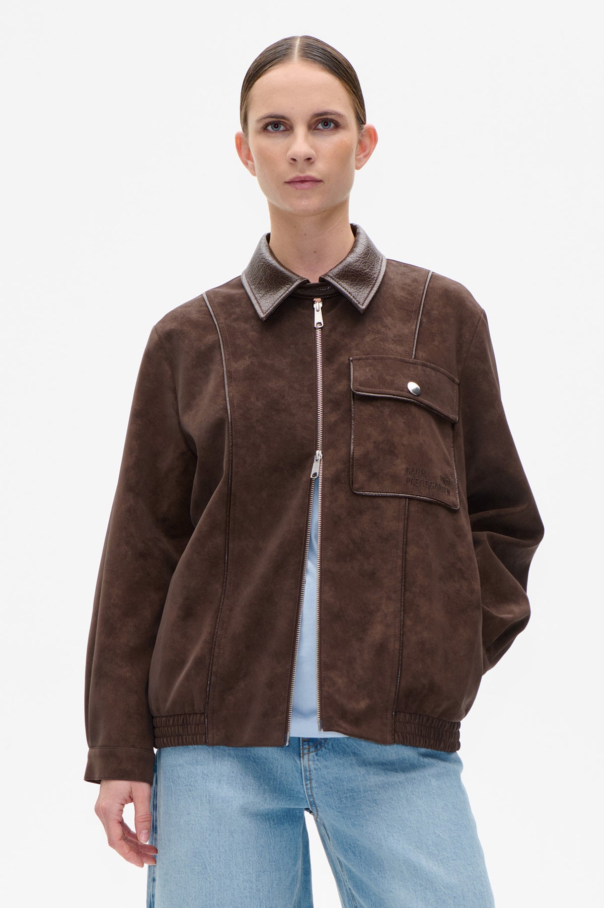 Front view of a model wearing a dark brown jacket with a shiny pointed collar, zip fastening, and snap-button chest pocket.