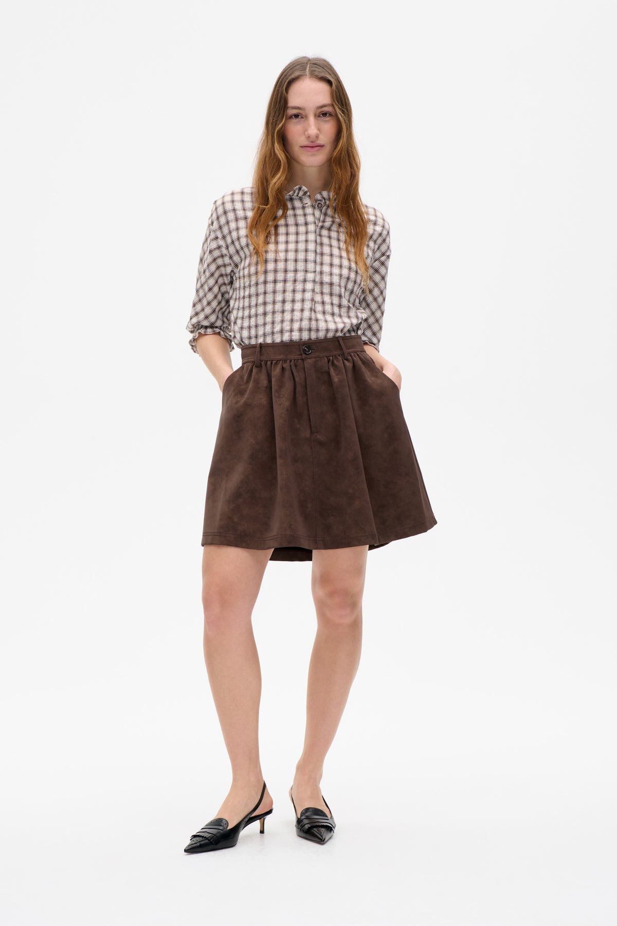 Front view of a model wearing a cream plaid blouse with ruffled collar and cuffs, a brown flared mini skirt and black pointed heels.