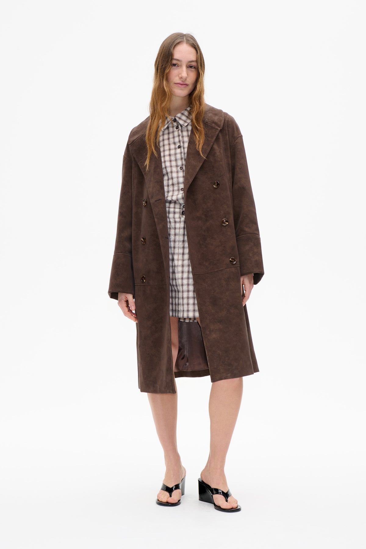 Model wearing a long brown double-breasted coat over a cream plaid shirt dress, styled with black heeled sandals against a white backdrop.
