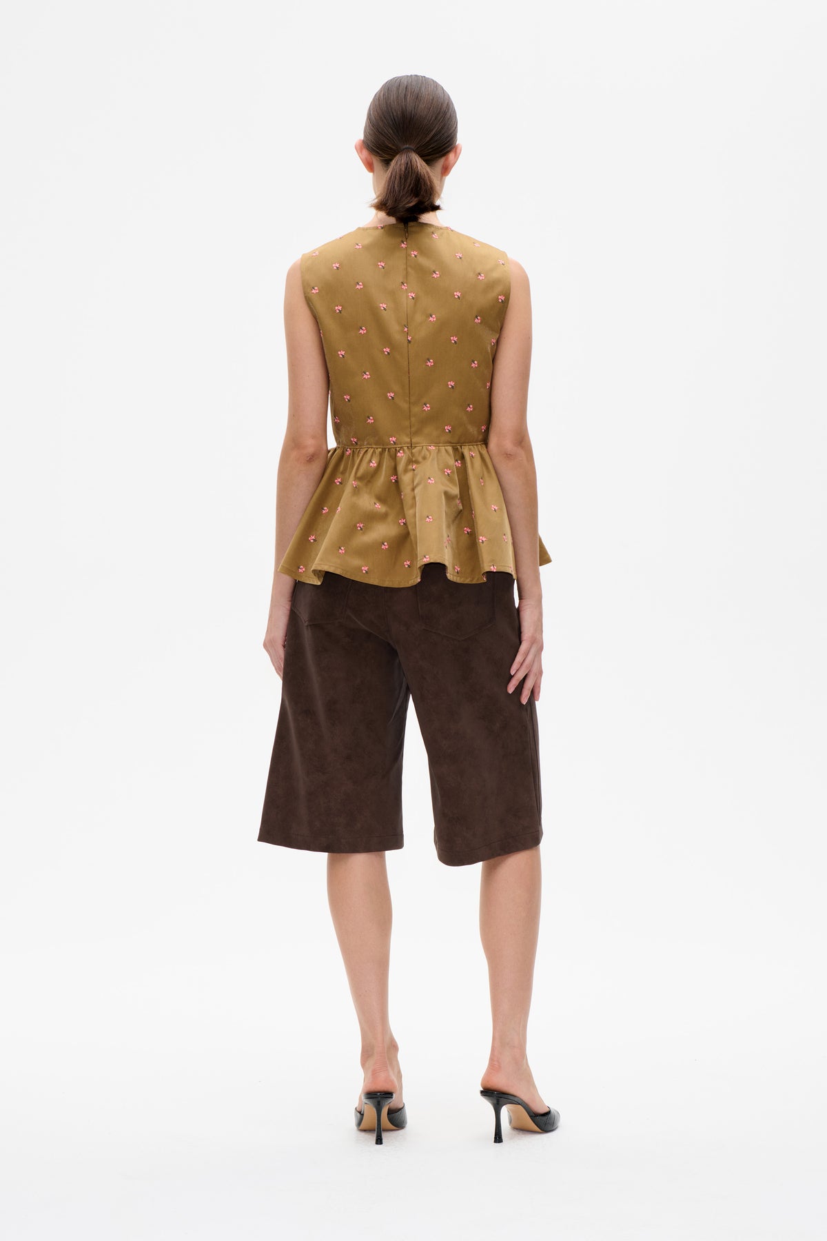 Rückenansicht eines Models in einem ockerfarbenen ärmellosen Top mit kleinen rosa Blüten und gerafftem Peplum, kombiniert mit einer dunkelbraunen, gekürzten Hose.