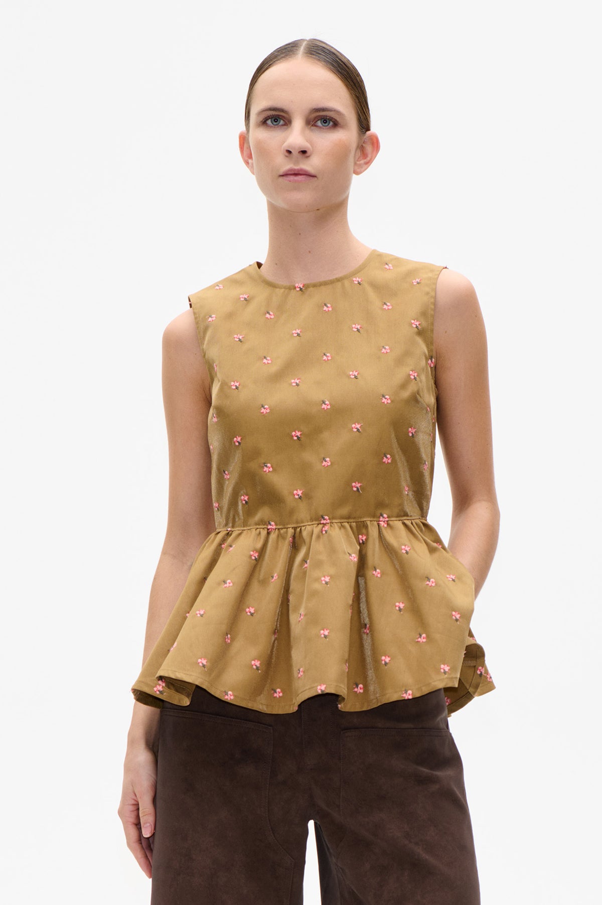 Model wearing a mustard-brown sleeveless top with scattered pink flowers and a gathered peplum hem, paired with dark brown trousers.