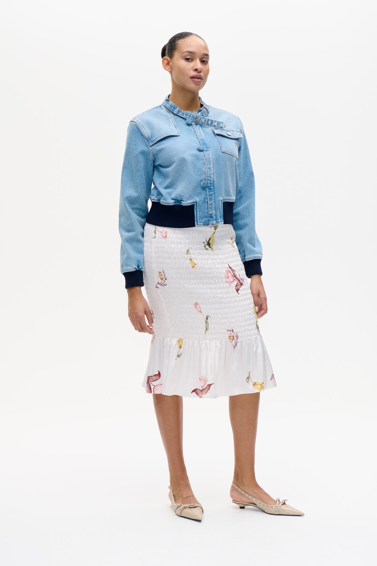 Model wearing a light blue denim jacket with a buckled eyelet collar strap and navy ribbed trim, paired with a white floral smocked dress.