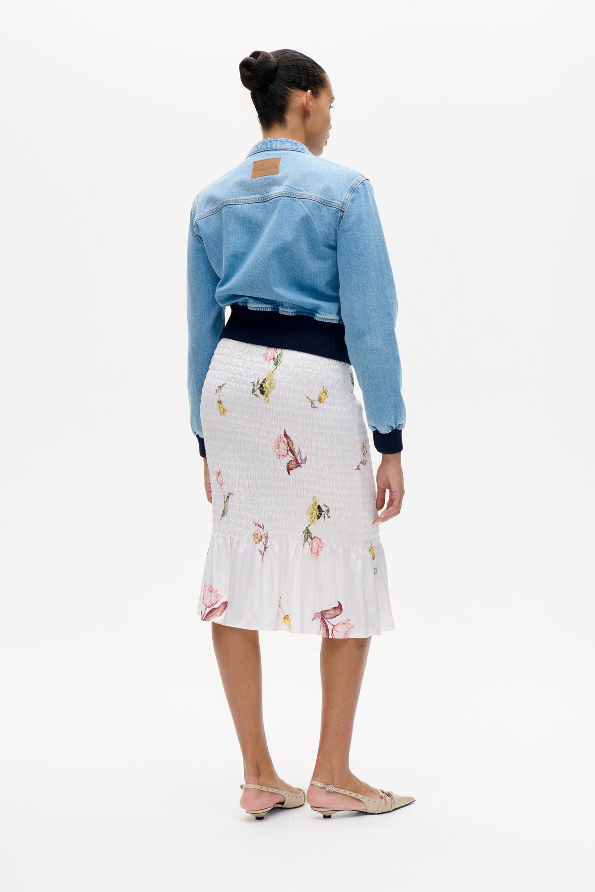Back view of a model wearing a light blue denim jacket with a tan logo patch and a white smocked floral skirt.