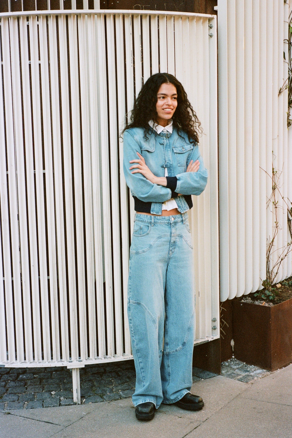 Campaign portrait of a model in a light blue denim jacket with dark cuffs and matching wide-leg jeans, standing before a white metal gate.
