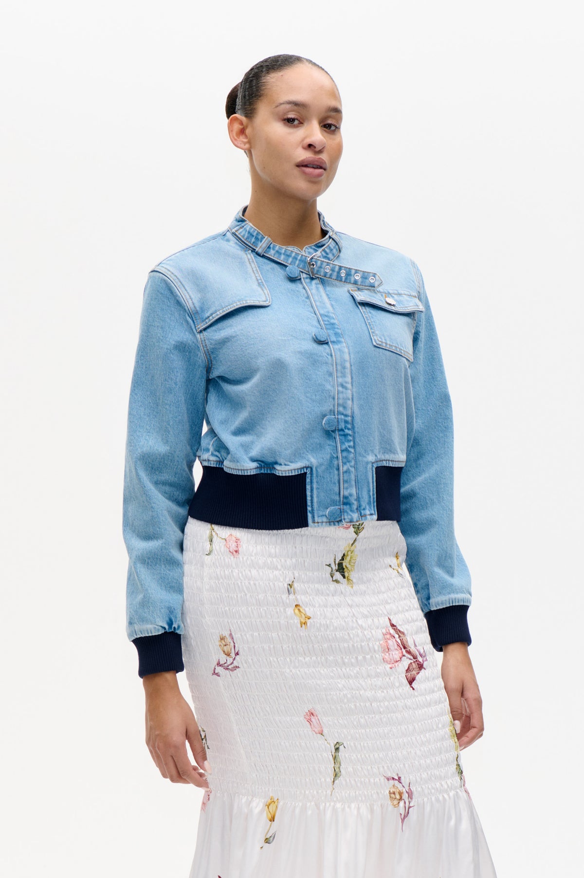 Model wearing a light blue denim jacket with a buckle collar and flap pockets over a white smocked floral dress.