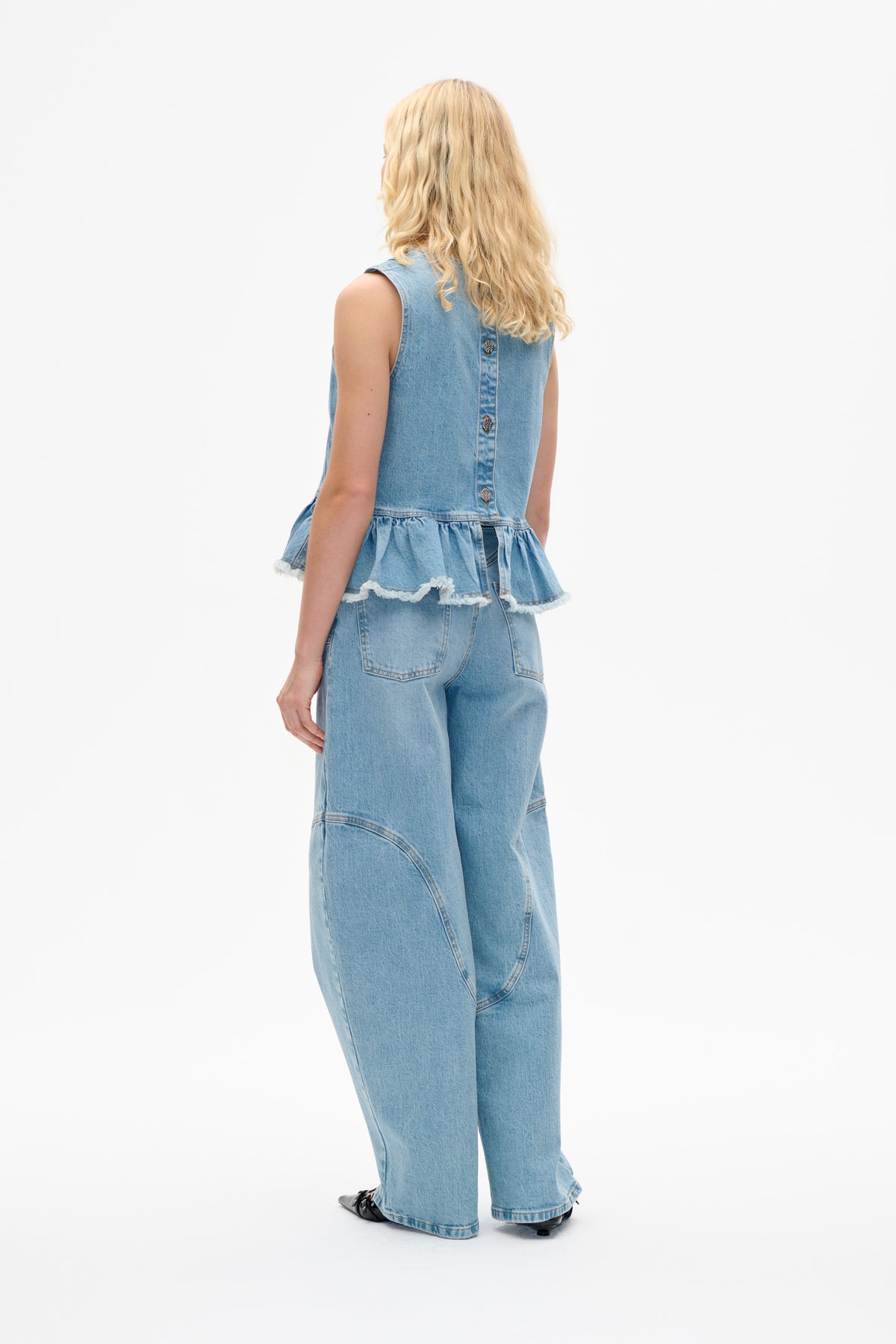 Back view of a model in a light-wash denim sleeveless top with a buttoned back and frayed peplum hem, paired with wide-leg jeans.
