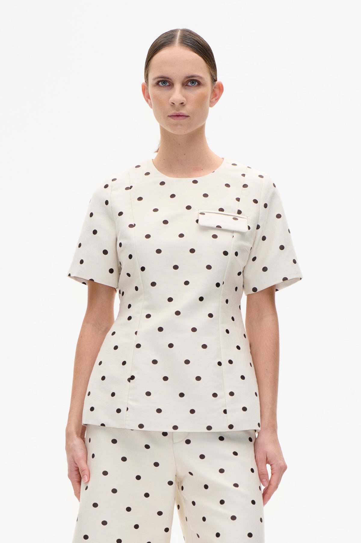 Front view of a cream short-sleeved top with dark polka dots and a chest pocket, paired with matching trousers.