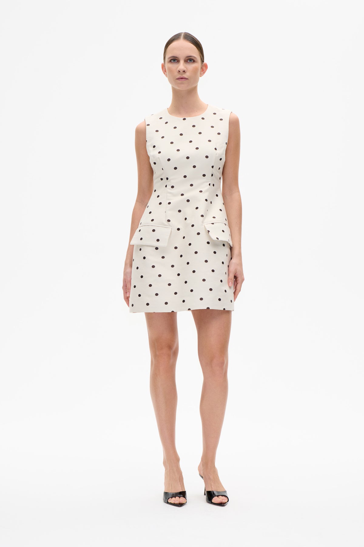 Front view of a sleeveless ivory mini dress with black polka dots and two large patch pockets.
